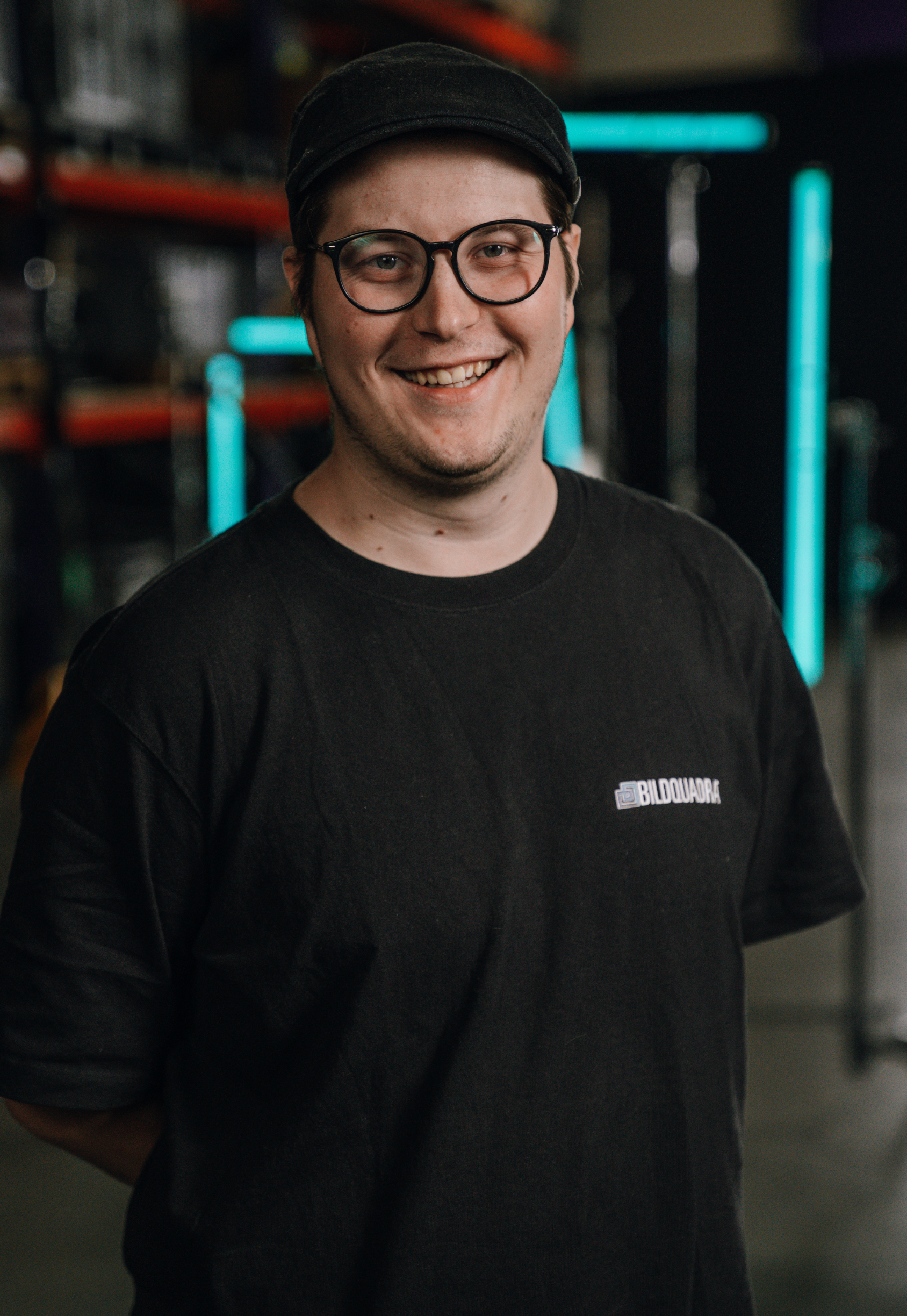 A young man with glasses and a black cap smiling at the camera, wearing a black T-shirt with a logo, in an industrial or workshop setting with shelves and neon lights.