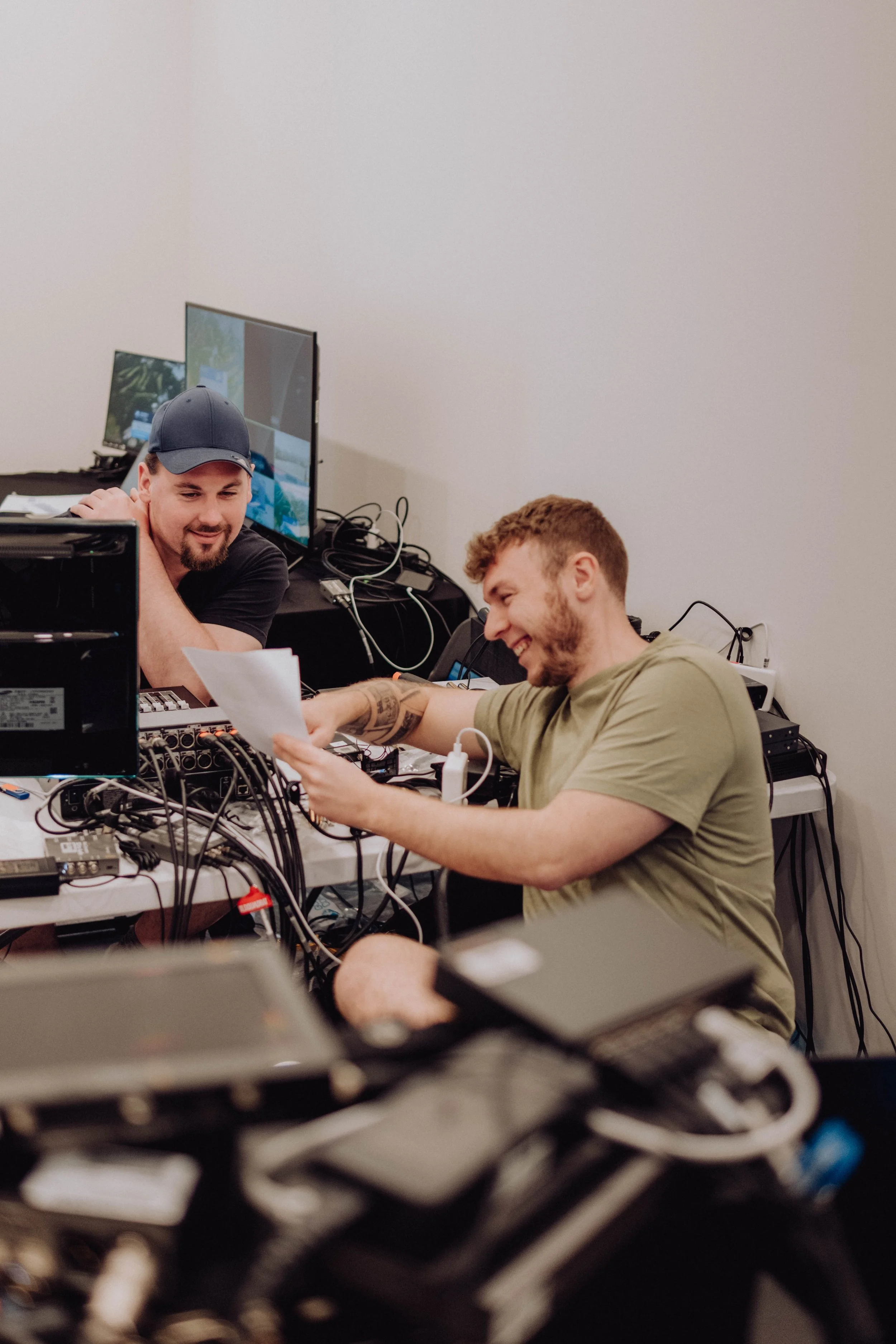 Two young men are laughing and smiling at a cluttered table with electronic equipment, wires, and monitors in a room with plain white walls.
