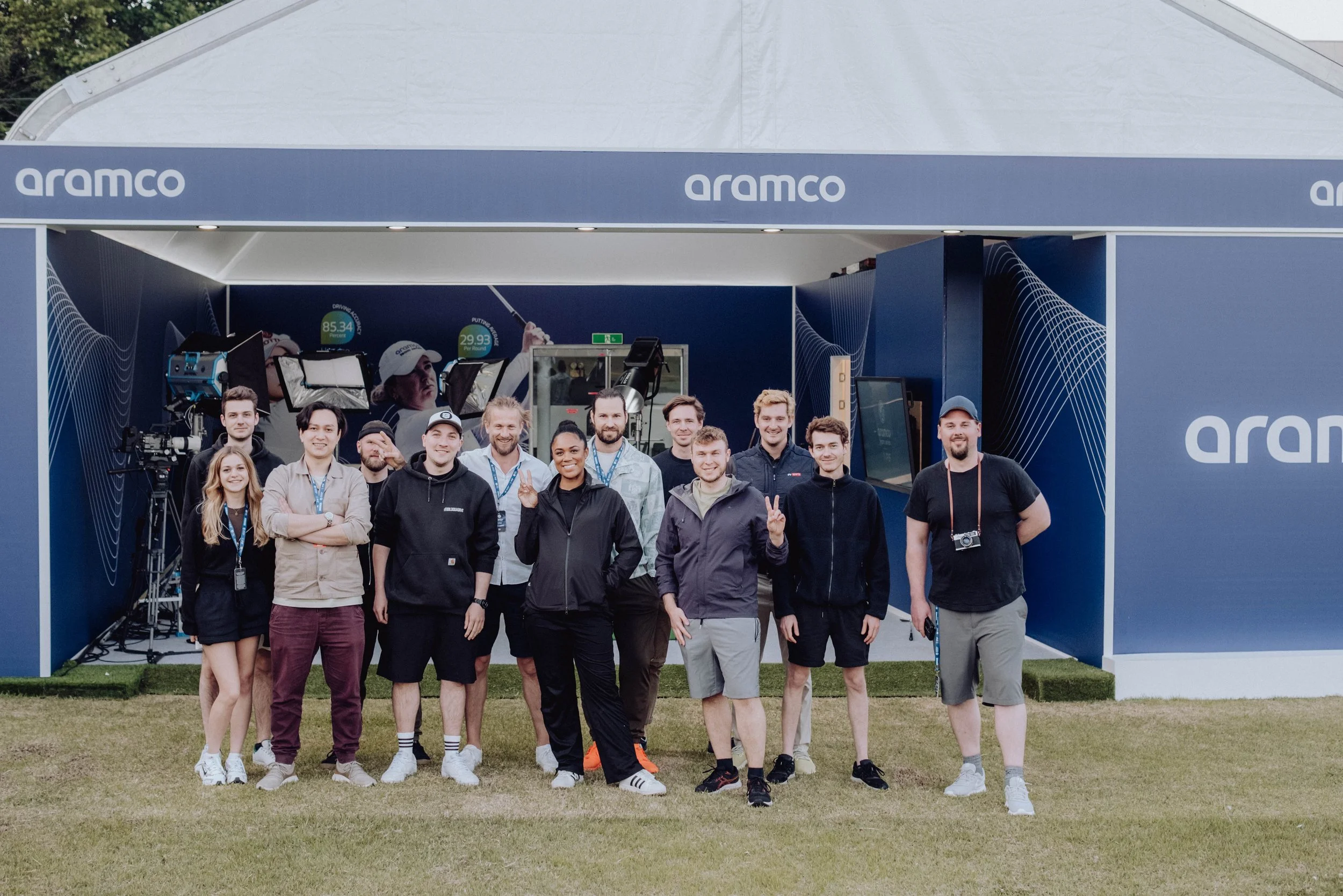Group of people standing in front of a blue Aramco tent with cameras and equipment behind them, outdoors on a grassy area.