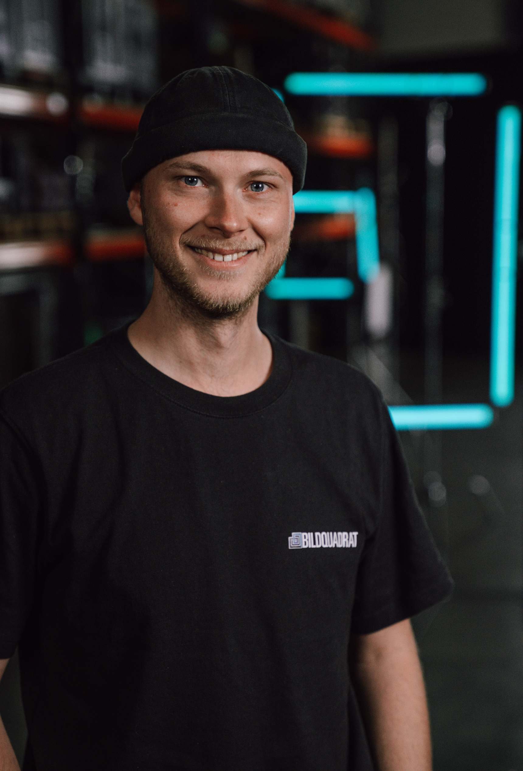A smiling young man wearing a black beanie and a black t-shirt with a logo, standing in front of a dark background with neon blue lights.
