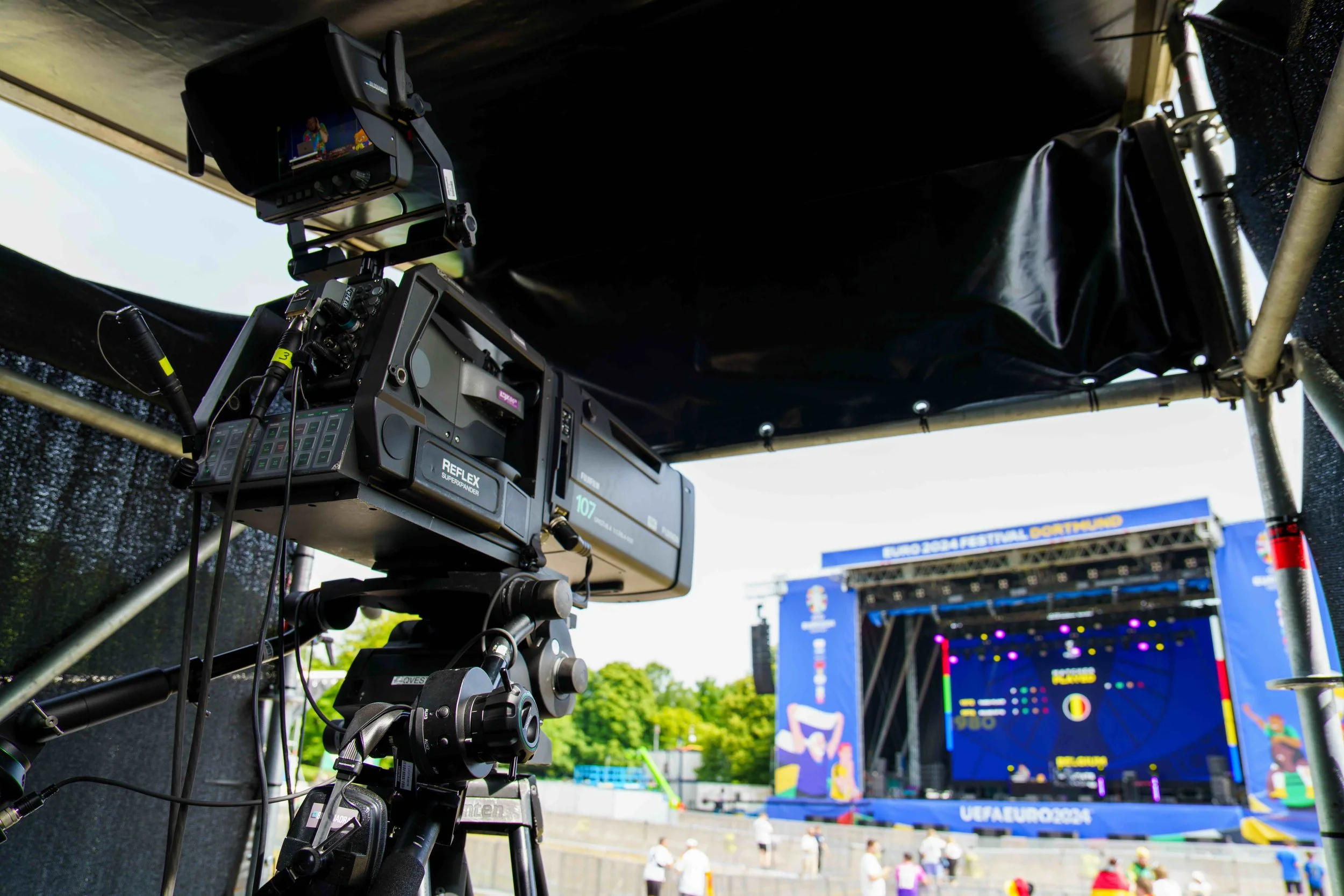 Camera equipment on a tripod set up to record a stage at an outdoor event during daylight, with a large screen displaying graphics and text in the background.