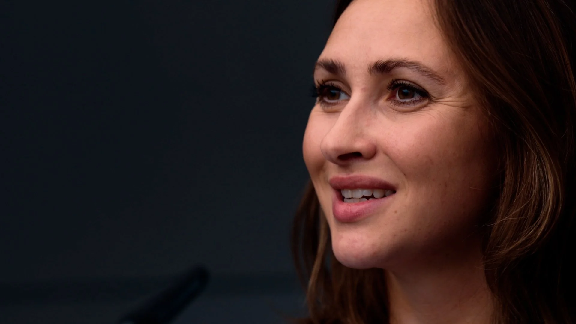 Close-up of a woman's face with brown hair, smiling and looking slightly to her right, against a dark background.