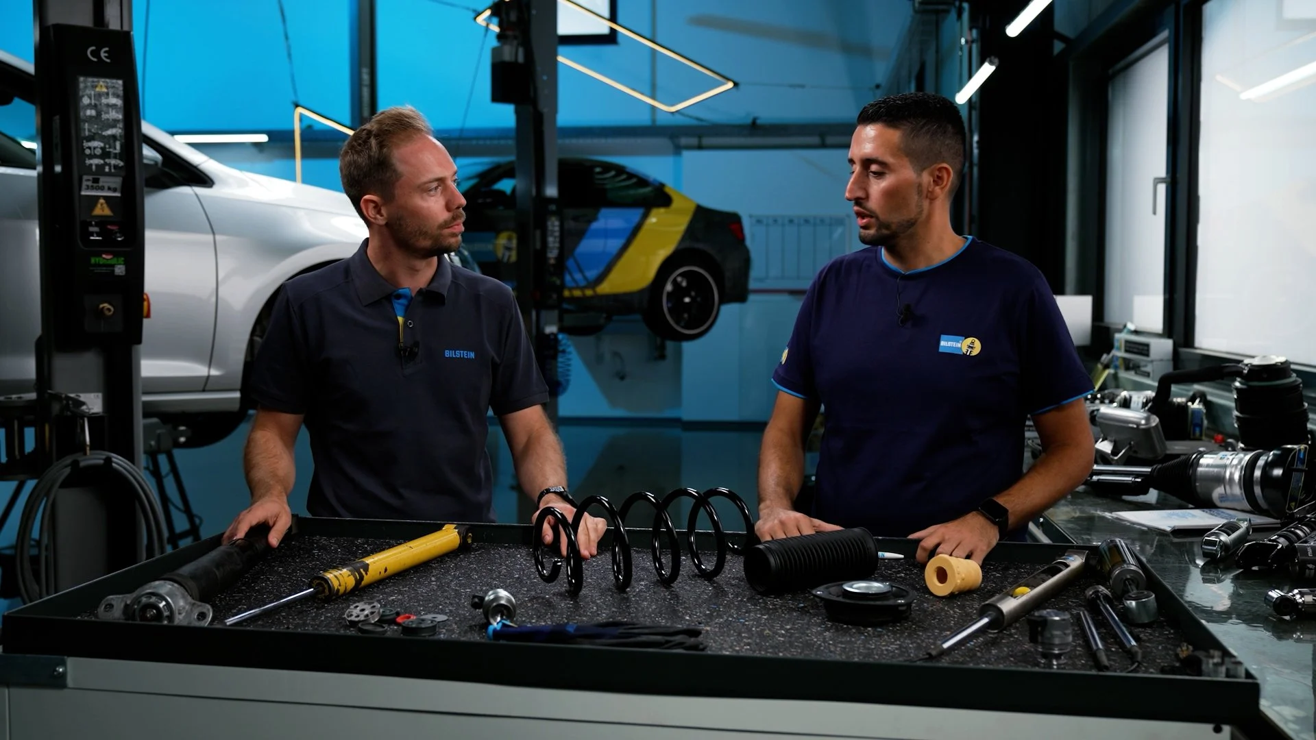 Two mechanics are having a discussion at a workbench with car suspension parts, in a modern auto repair shop with cars on lifts in the background.