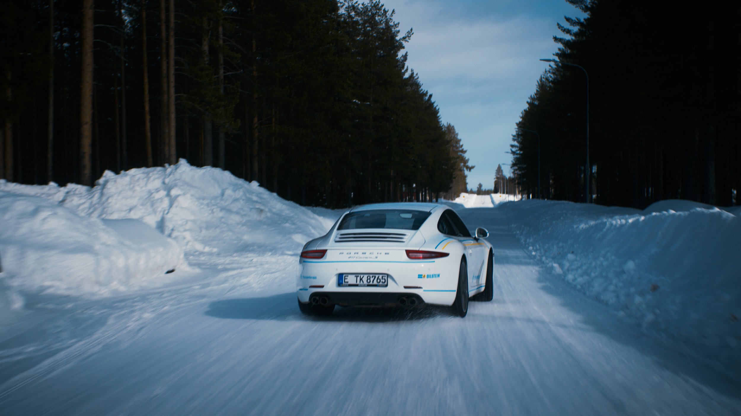 A white Porsche Taycan driving on a snowy road surrounded by snow banks and forested trees during daytime.