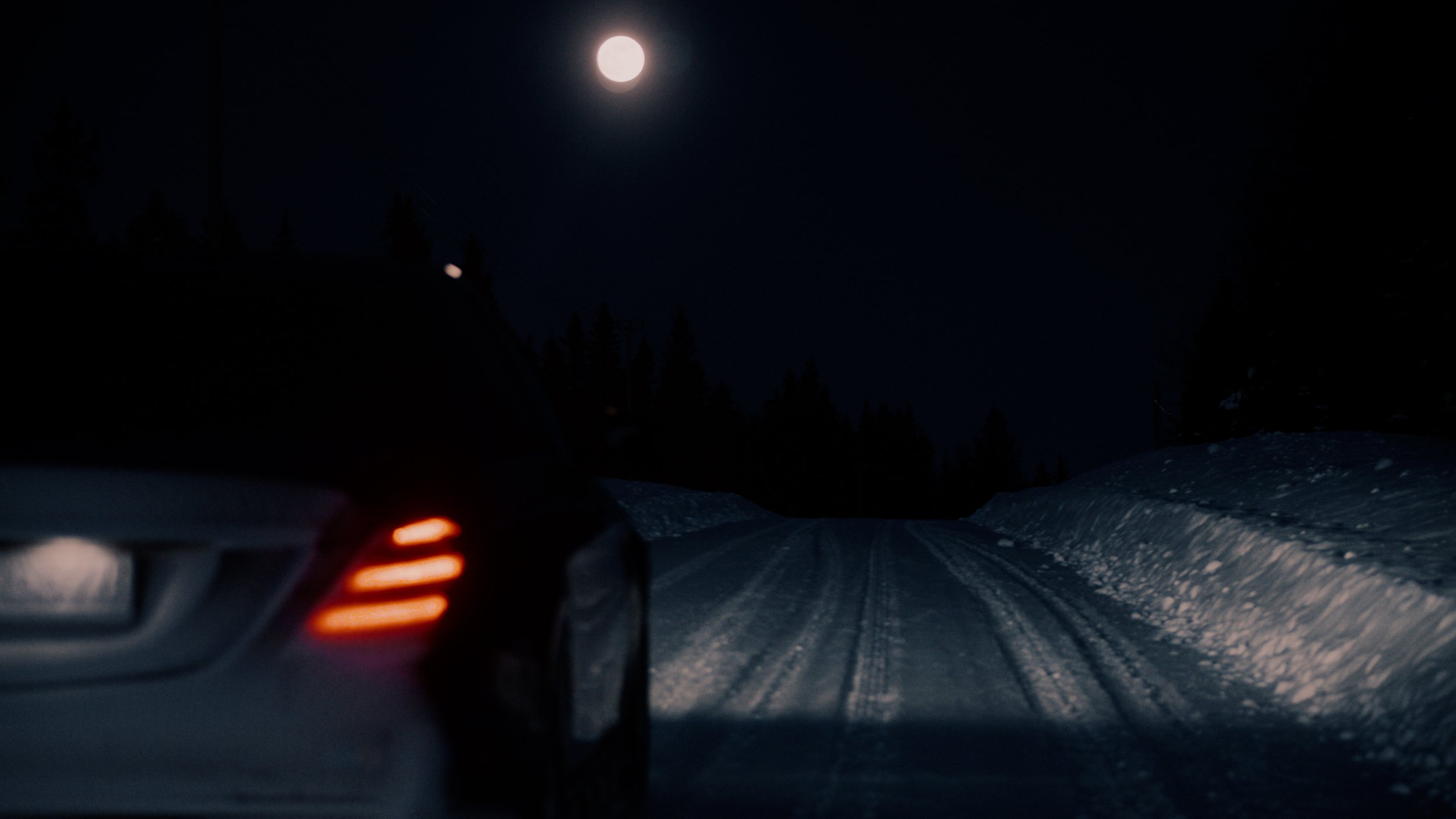Nighttime scene showing a snow-covered road flanked by snow banks, with a bright full moon in the sky, and trees in the background. Part of a vehicle with illuminated taillights is visible on the left side.