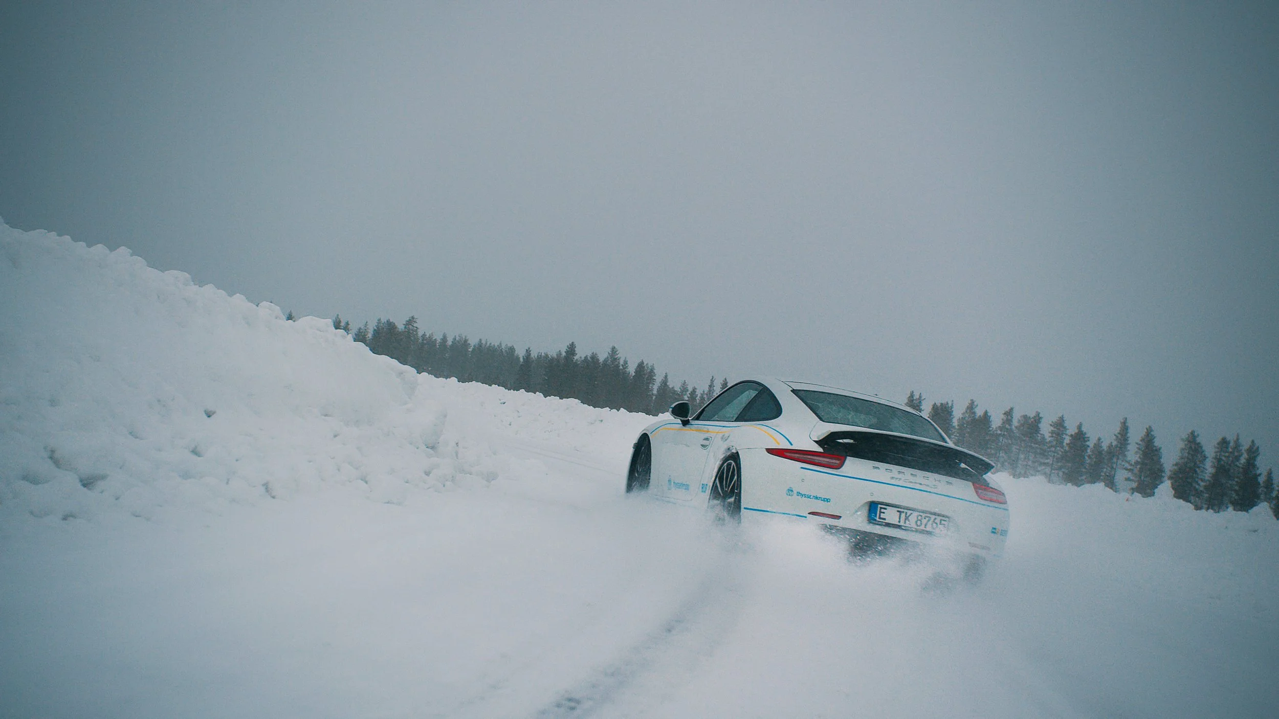 A white Porsche sports car driving through a snow-covered landscape with snowbanks on the side, leaving tire tracks in the snow.