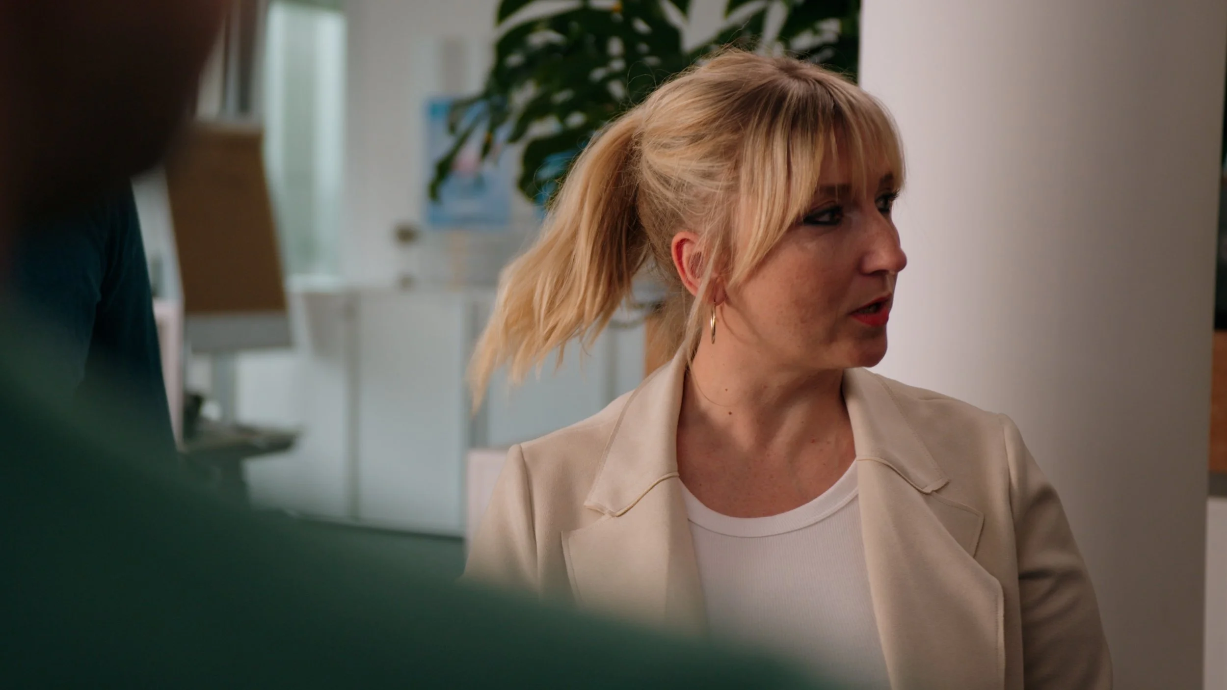 A blond woman with a ponytail and hoop earrings, wearing a light-colored blazer and white top, is speaking or listening in an indoor setting with plants and office furniture in the background.