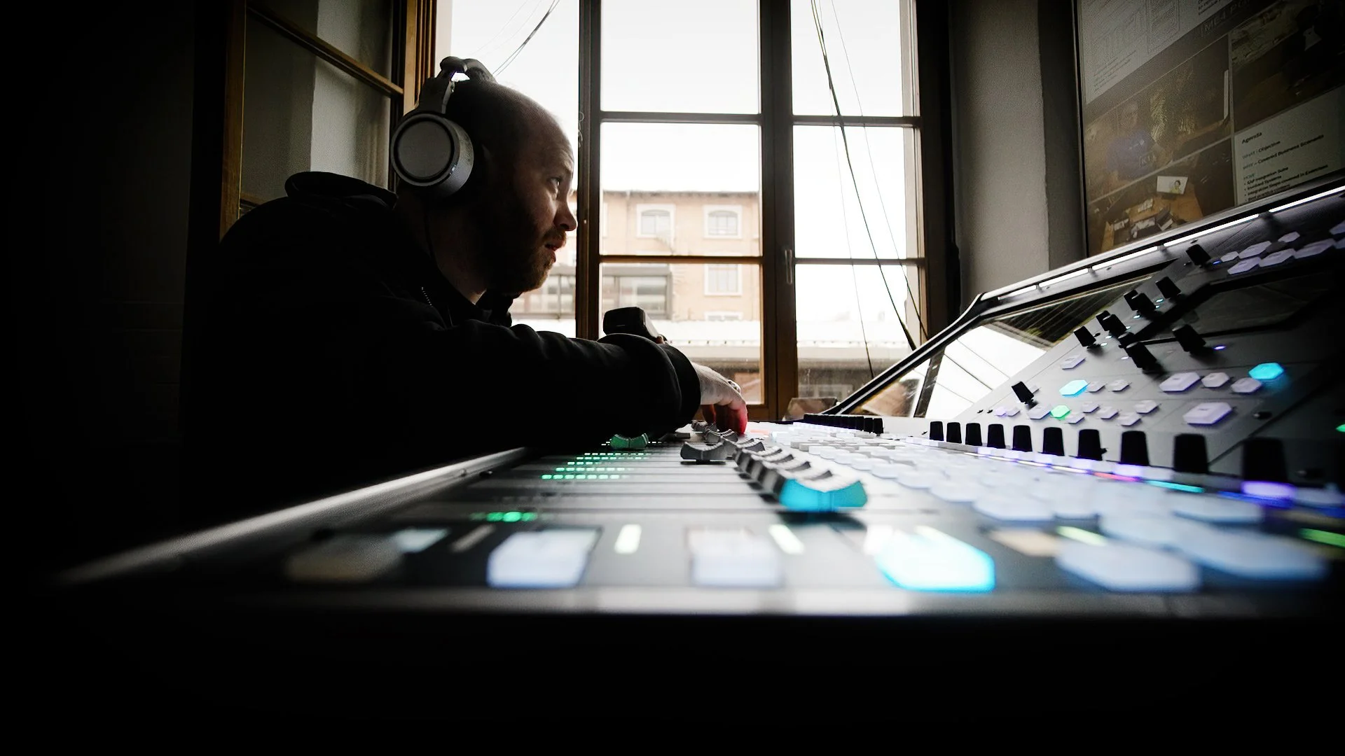 A man wearing headphones works on a music production console with colorful lights, sitting near a window with an urban view.