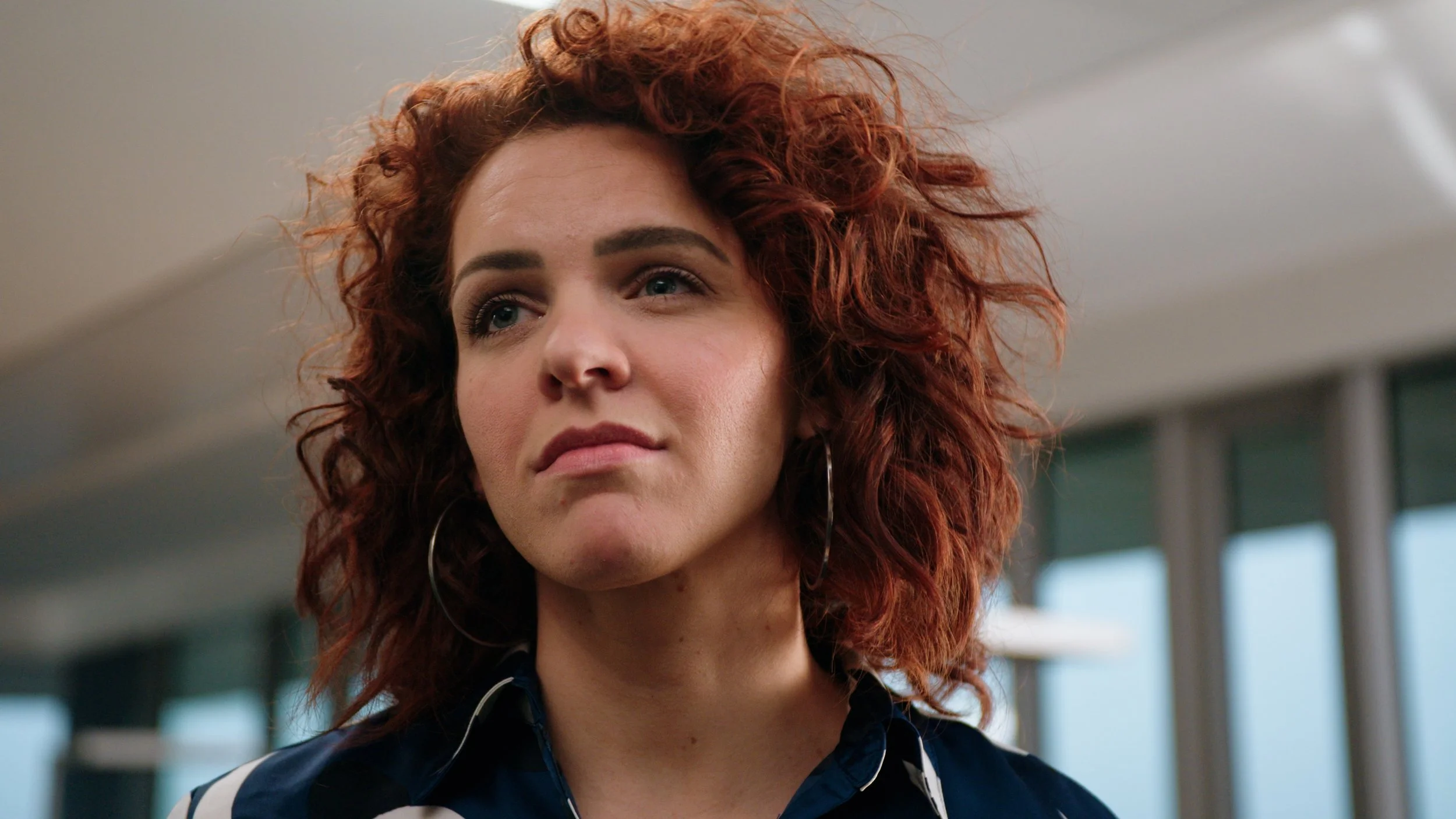 A woman with curly red hair, wearing hoop earrings, looking slightly displeased or skeptical.
