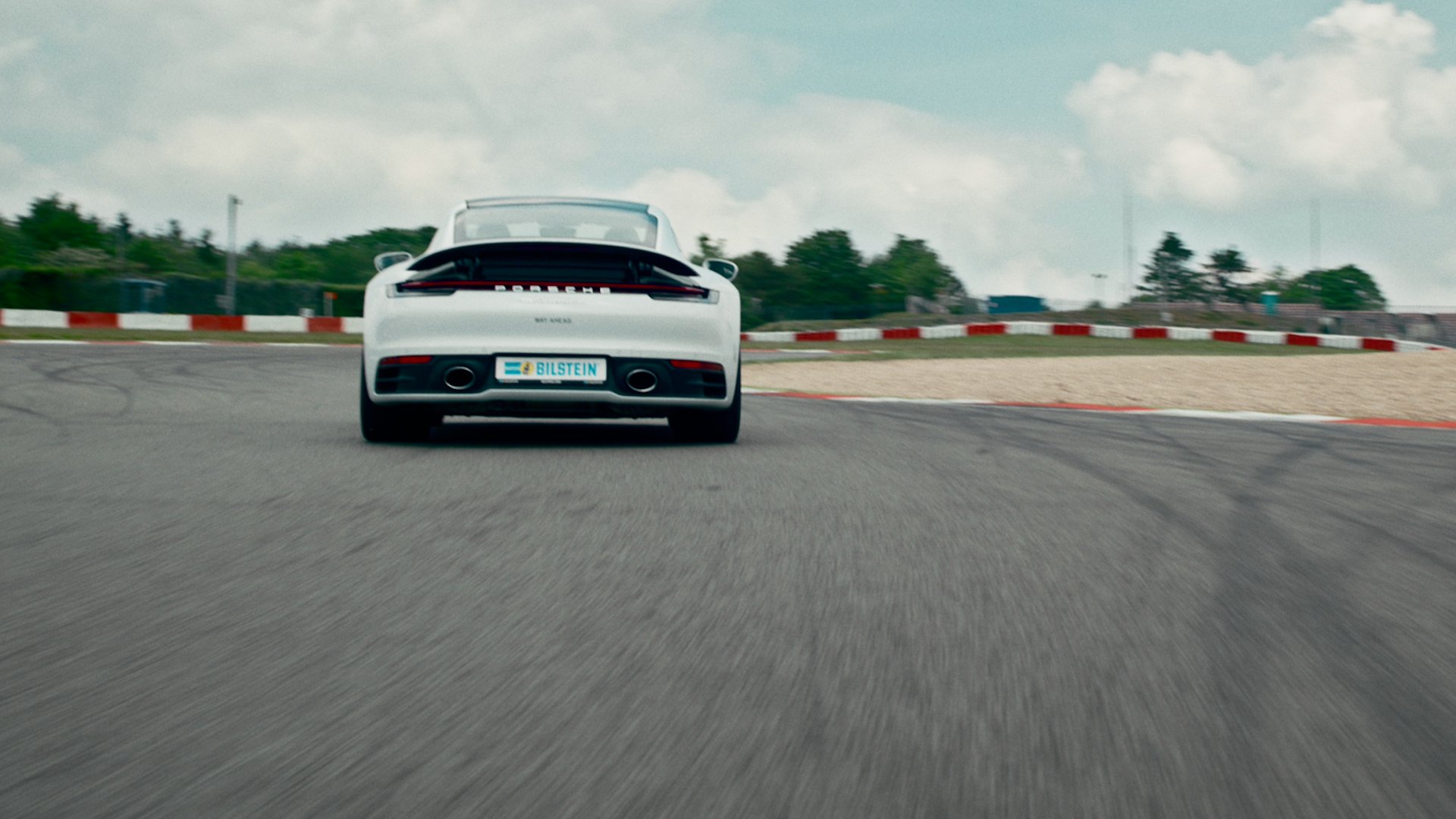 White Porsche sports car driving on a race track with red and white curbs and green trees in the background.
