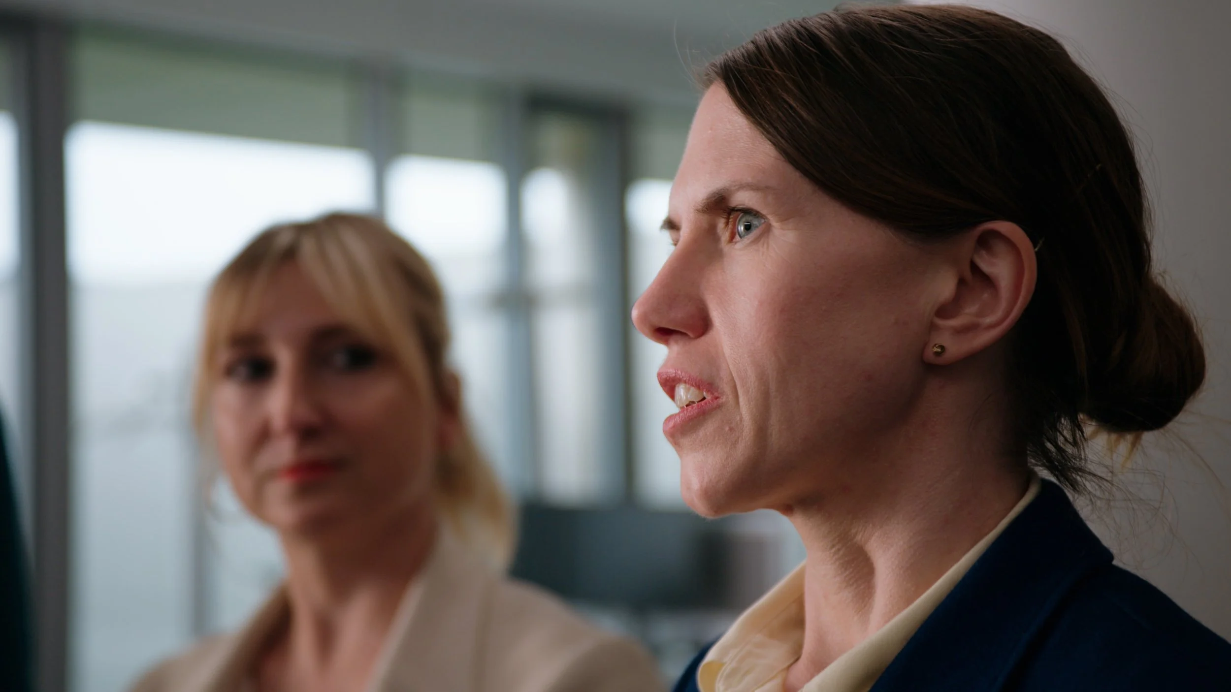 Two women in a professional setting, one with dark hair in a bun and the other with blonde hair, engaged in conversation against a background of large windows and natural light.