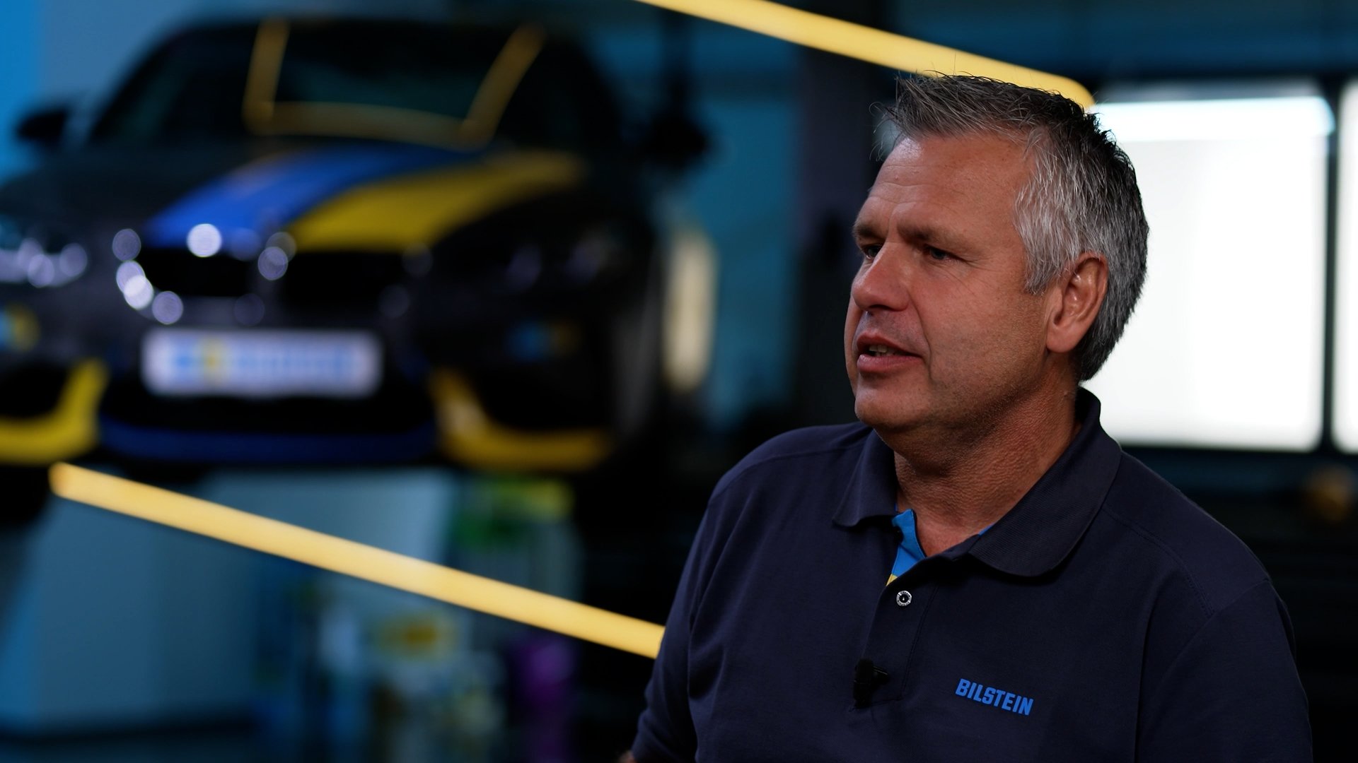A man with gray hair wash speaking indoors, with a black and yellow sports car in the background.