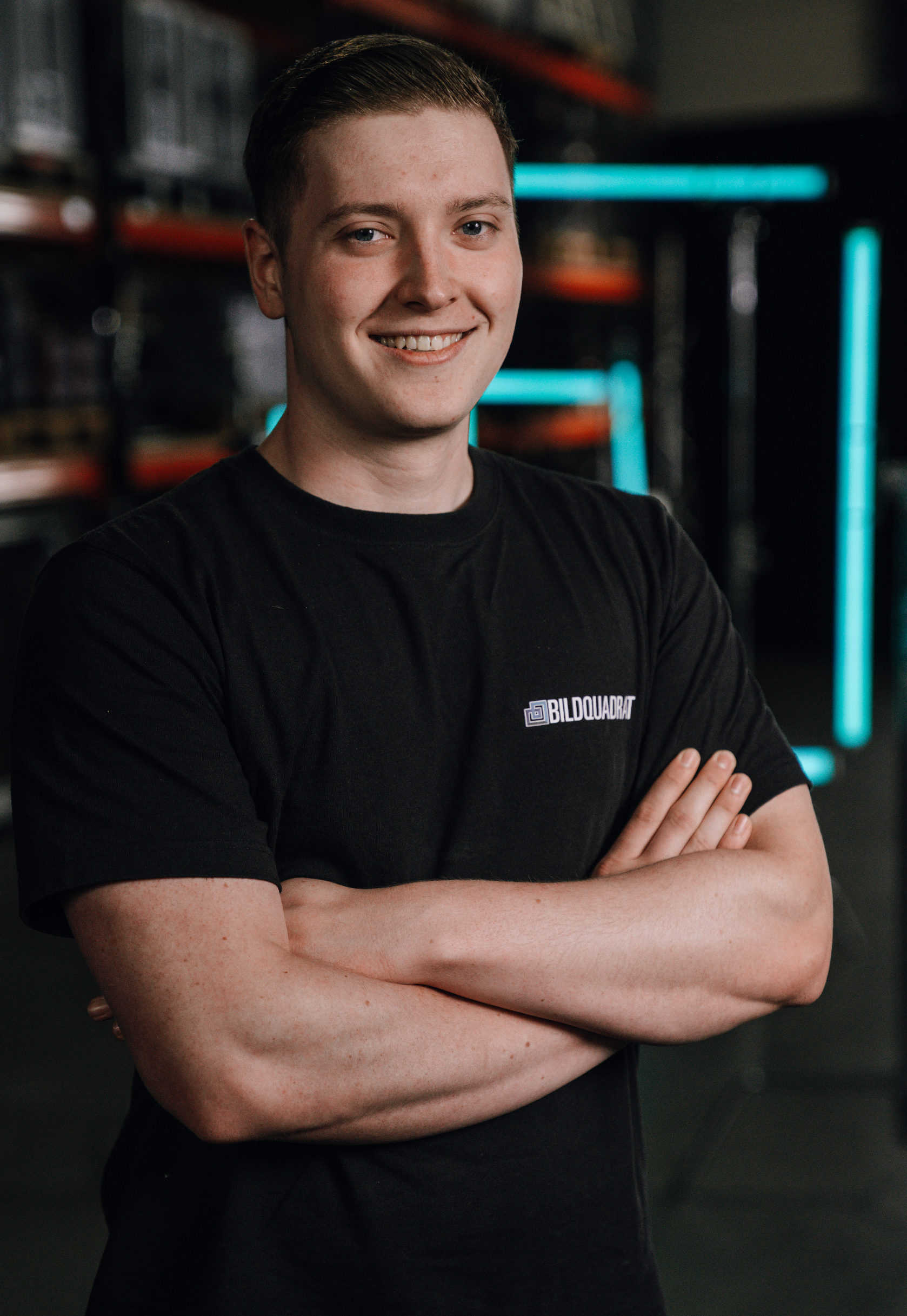 A young man with short brown hair and light skin, smiling with arms crossed, wearing a black T-shirt with a logo that says 'BILDQUADRAT,' standing in a dark industrial setting with neon blue lights in the background.