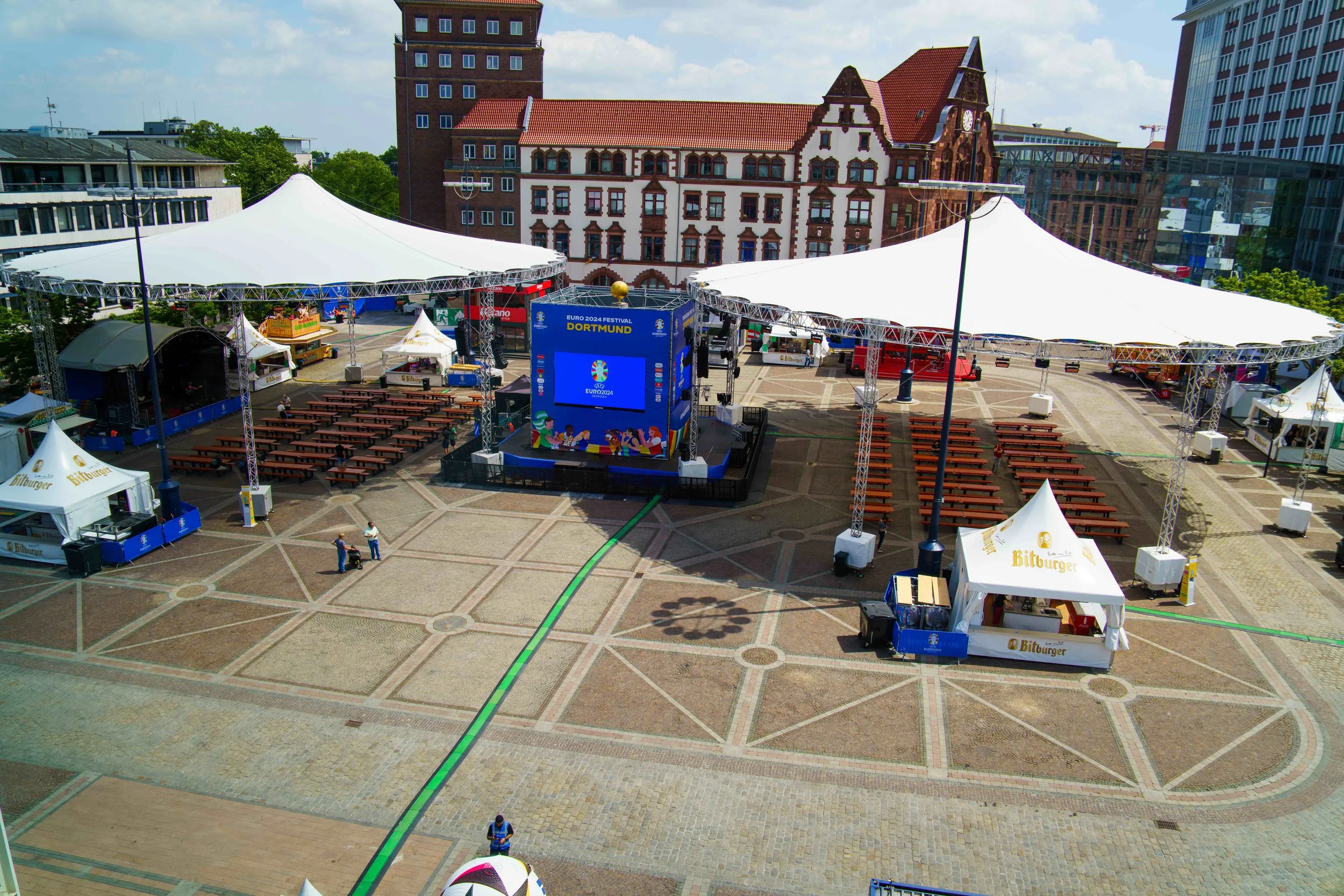 An outdoor event space set up with large white tents, wooden picnic tables, a stage with a large screen displaying "EURO 2024 Dortmund," and food tents with 