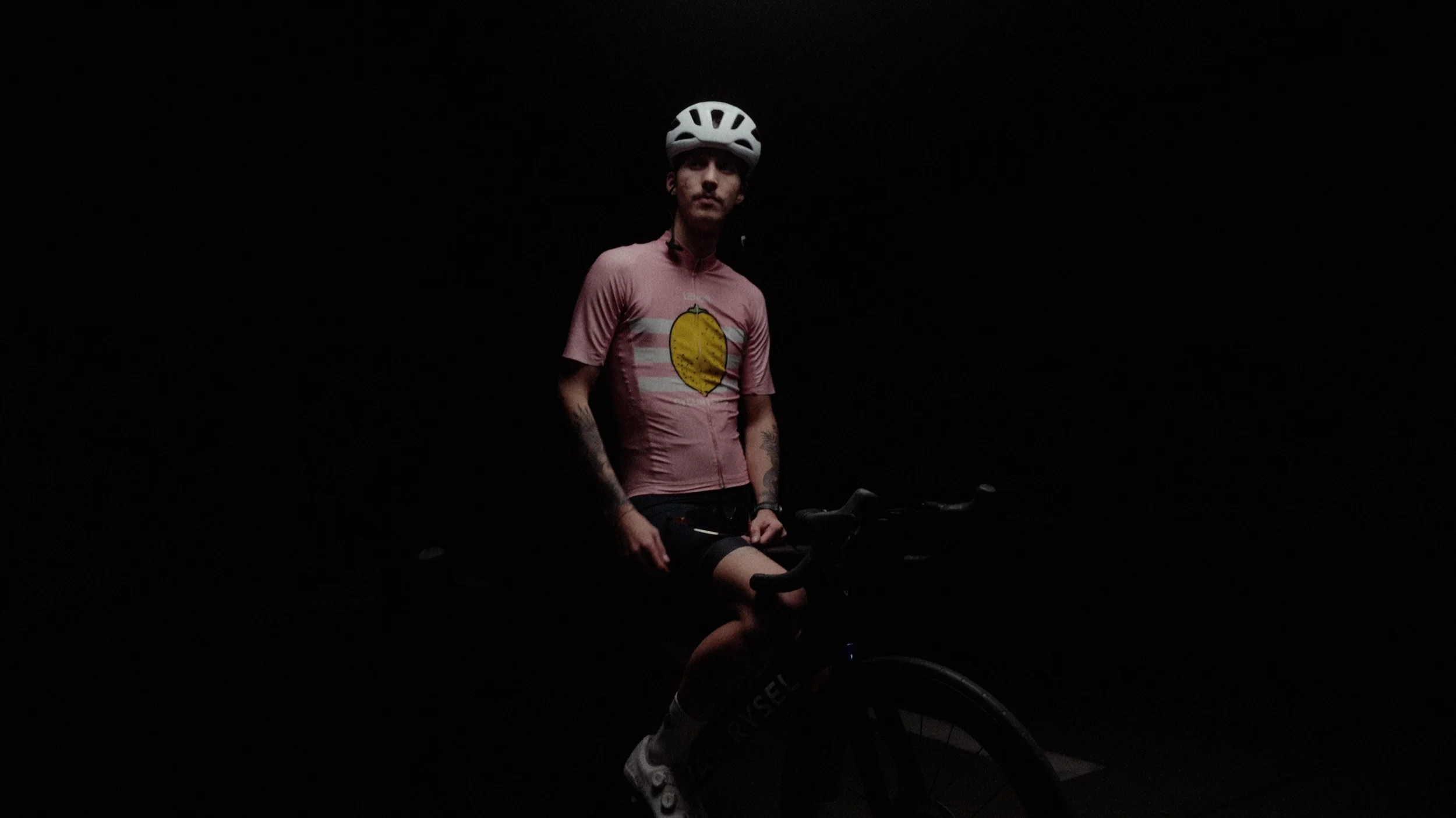 A cyclist in a pink jersey with a lemon graphic, wearing a white helmet, sitting on a black bicycle in a dark environment.