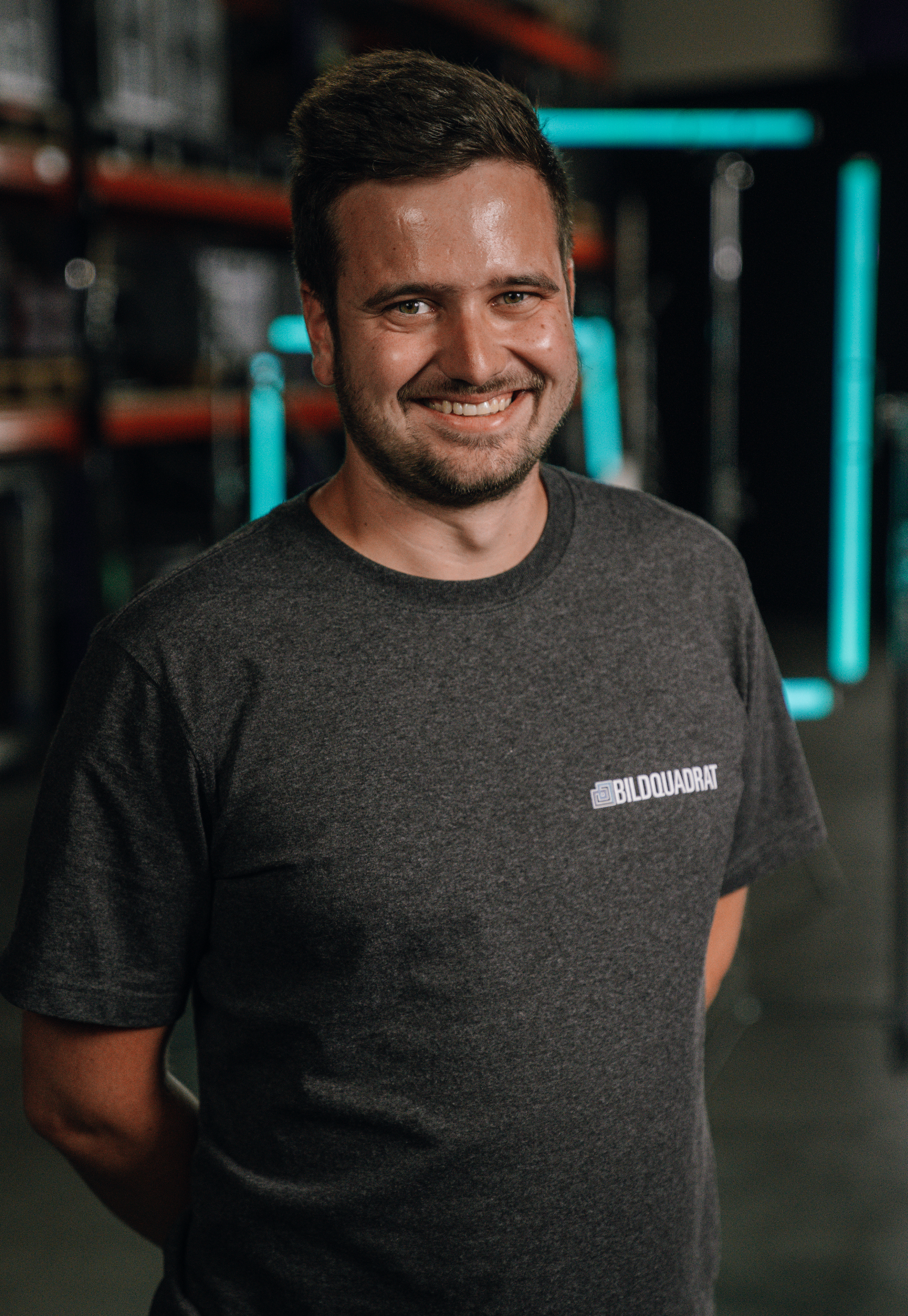 A man with short dark hair, beard, and smiling face, wearing a gray T-shirt with the logo 'BILDQUADRAT,' standing in an indoor gym with shelves and neon lights in the background.