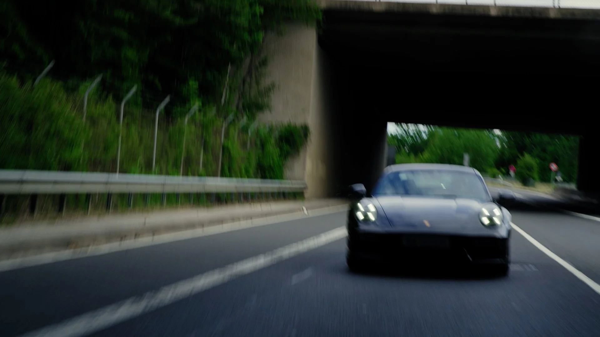 A black sports car, possibly a Porsche, driving on a highway under a bridge with green trees on the side.