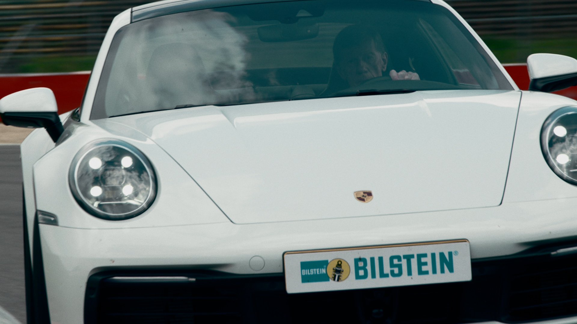 Front view of a white Porsche sports car with black accents, around a race track, with a person visible through the windshield.