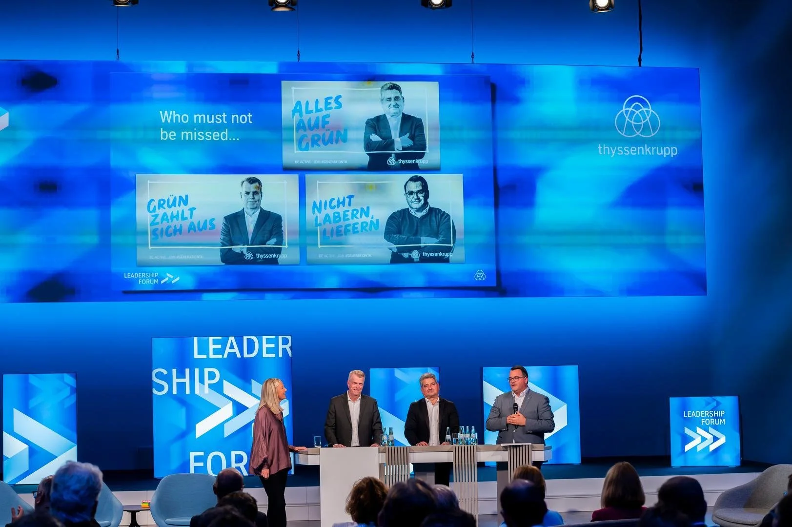 Four people on stage at a leadership forum conference, with two men and one woman standing and one man speaking into a microphone. The stage backdrop has blue panels displaying the words 'LEADERSHIP FORUM' and large screens showing images and message
