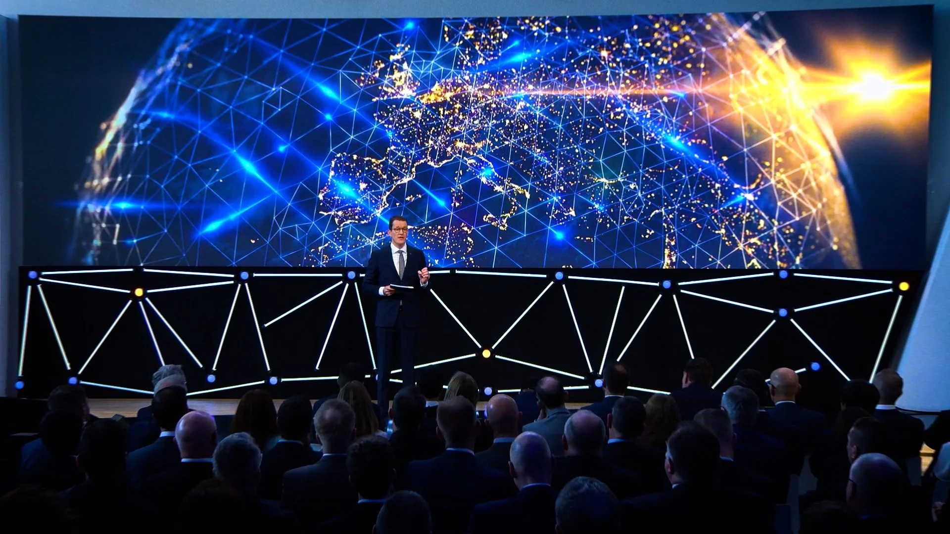 A man in a suit giving a presentation on stage with a large digital world map in the background and a networked globe of interconnected lines. Audience members are watching.