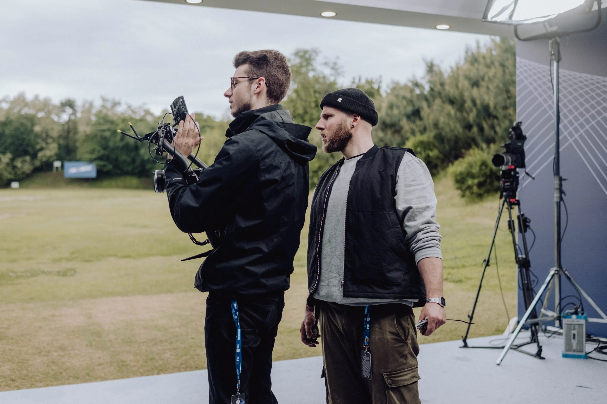 Two men working on a film set outdoors, one holding a camera and the other looking at the equipment, with a professional camera on a tripod and lighting setup nearby.