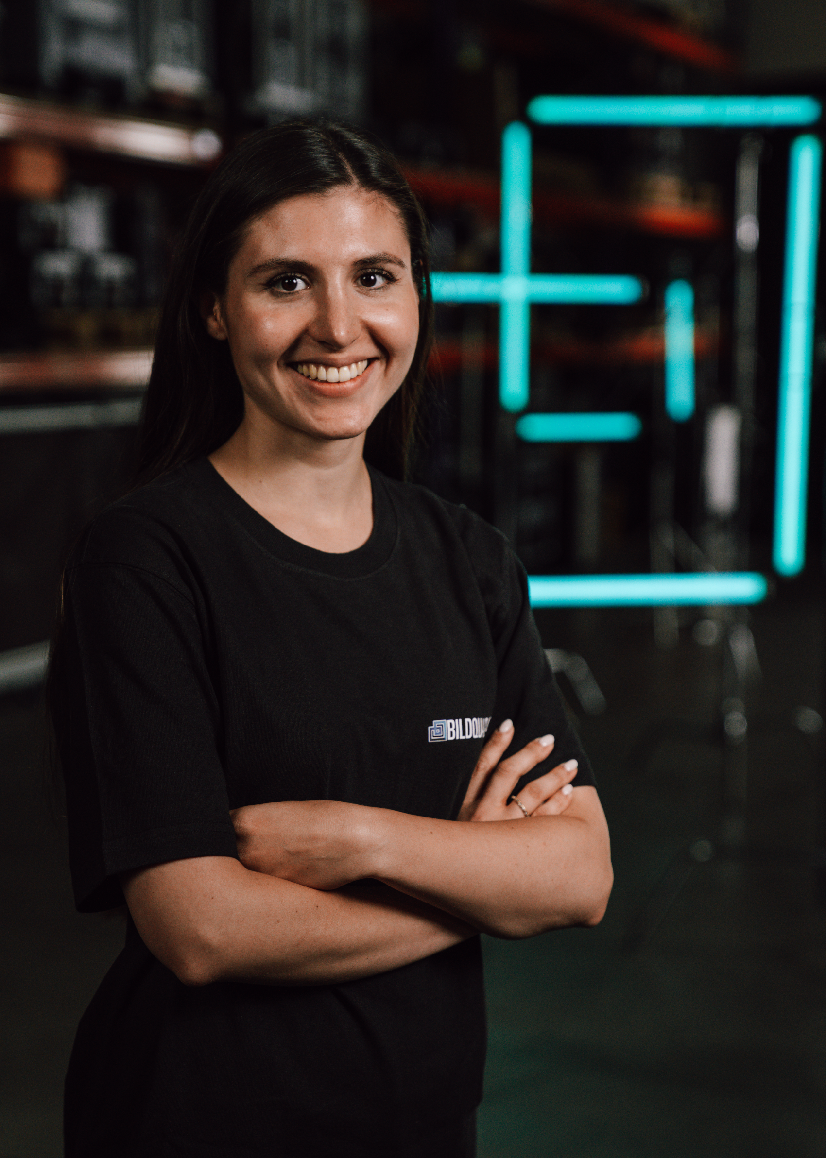 A young woman with long dark hair, smiling and crossing her arms, standing in a dimly lit room with neon blue geometric light art in the background.