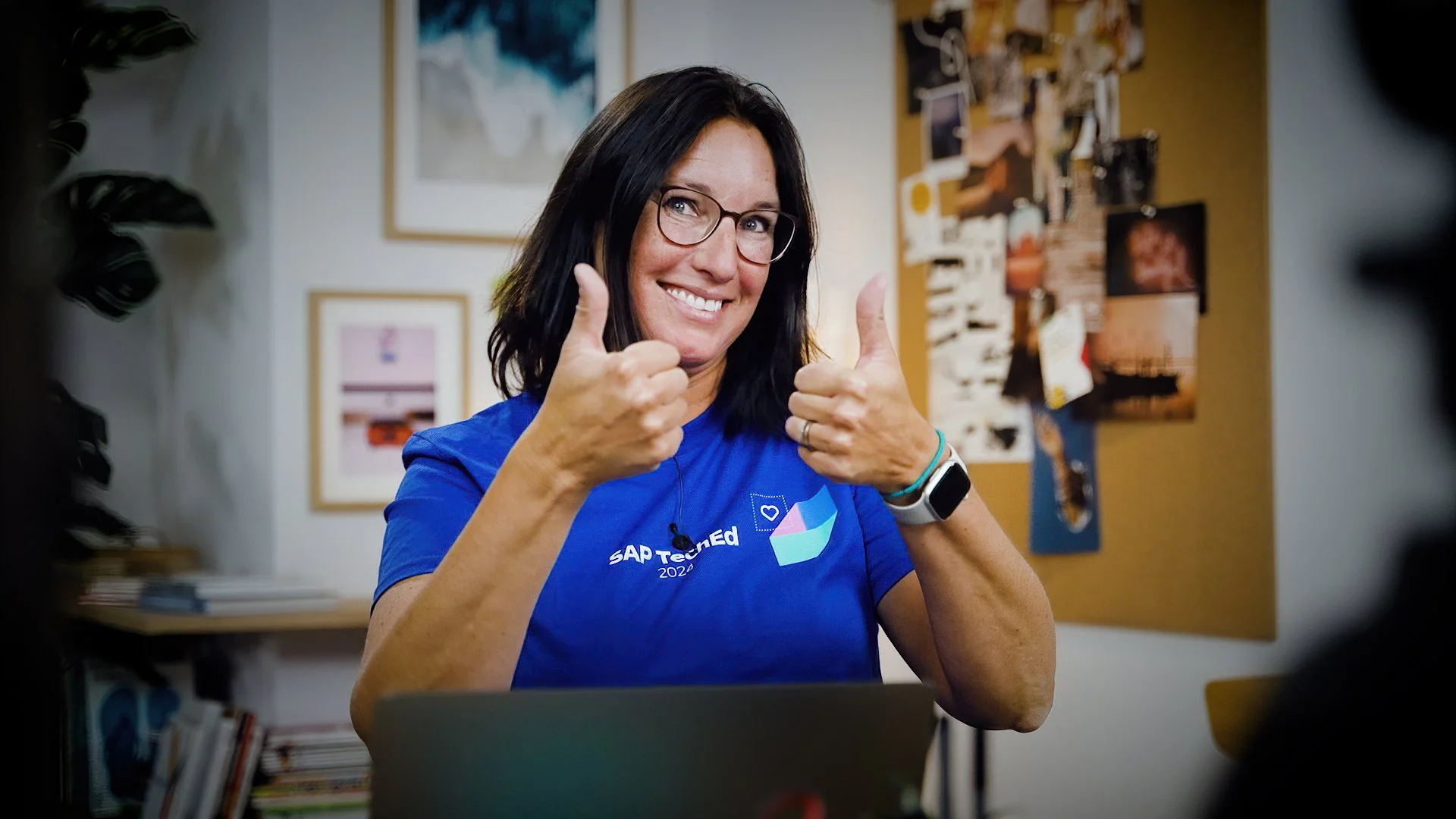 A woman with dark hair, glasses, and a blue T-shirt giving a thumbs-up and smiling while sitting at a desk with a laptop. The background includes framed pictures and a bulletin board.