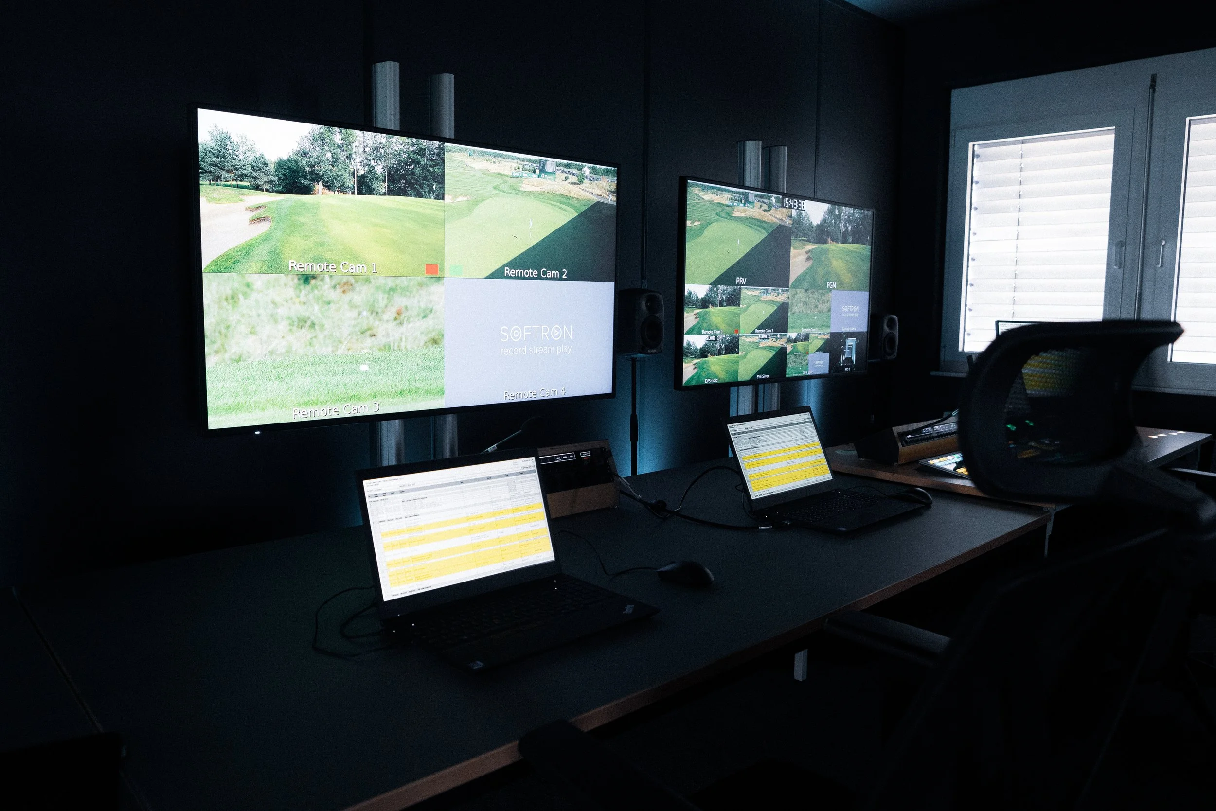 Golf simulation room with two large screens displaying golf courses and multiple smaller screens and laptops on a desk.