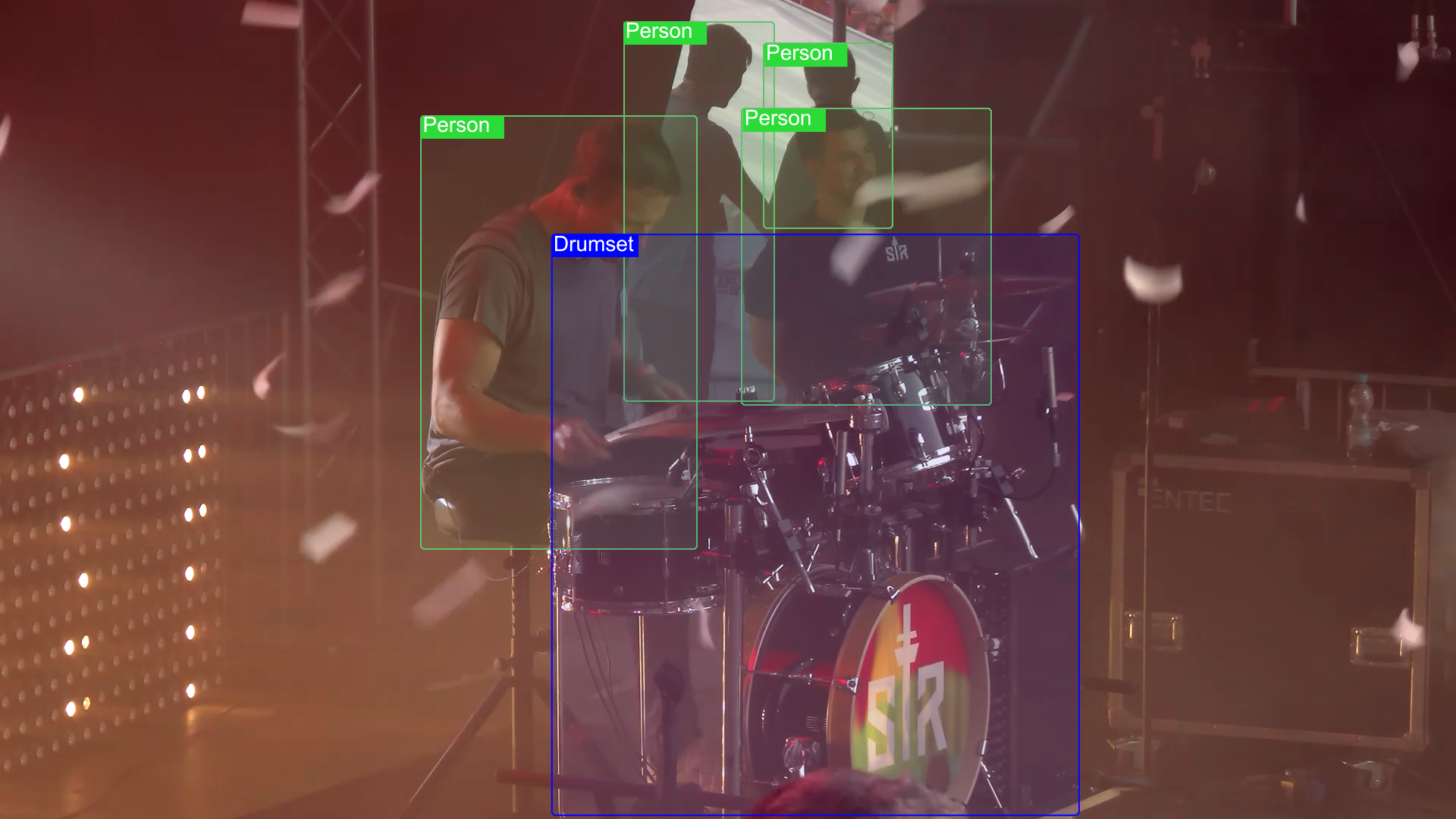 A group of four people on stage, with one playing a drum set, and the others standing around, with confetti falling, illuminated by stage lights.