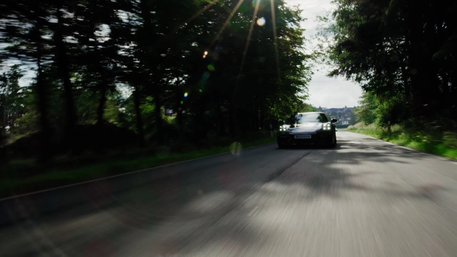 A black sports car driving down a rural road surrounded by trees, with sunlight creating lens flares in the image.