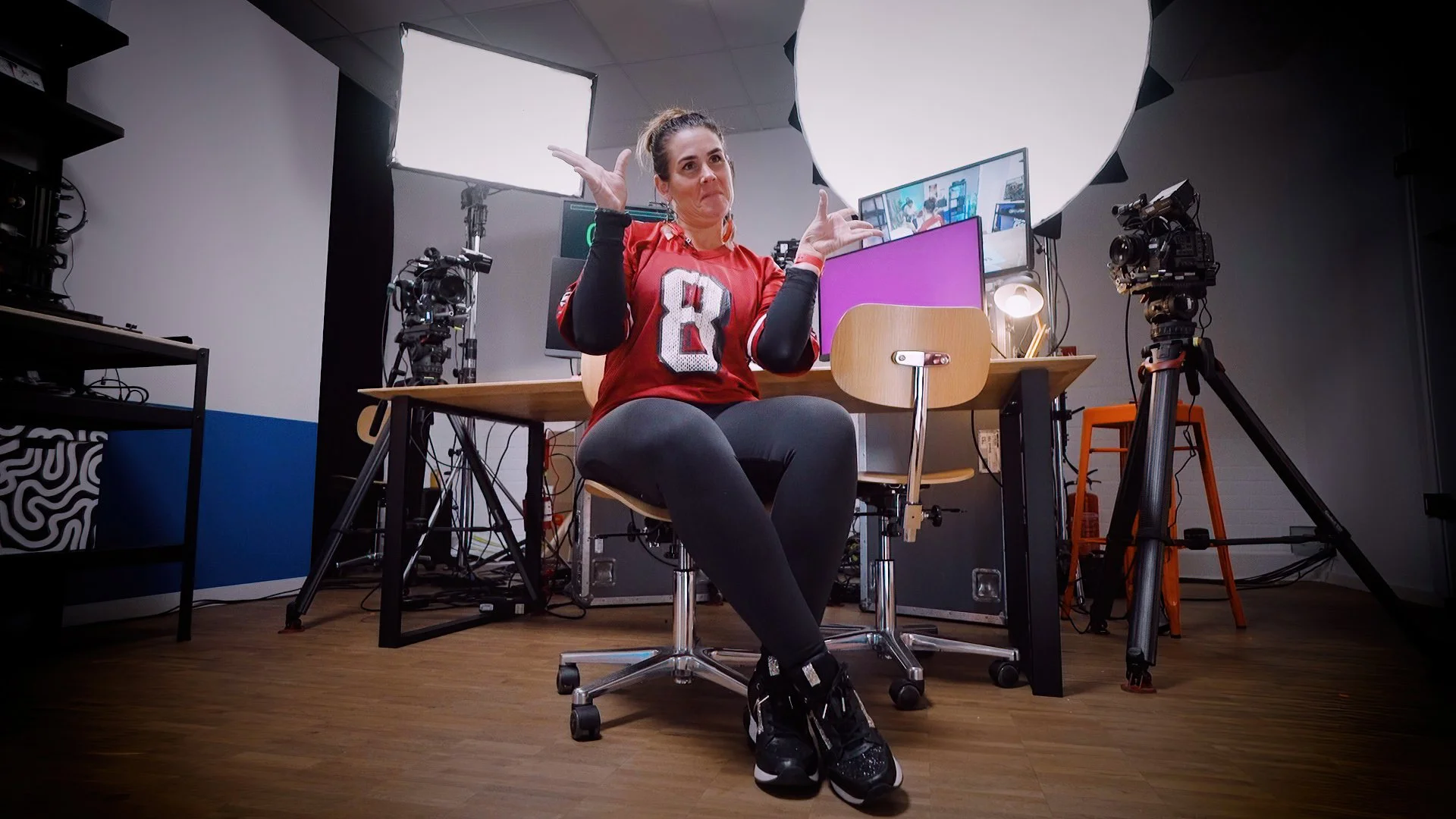 A woman sitting on a chair in a recording studio, gesturing with her hands, surrounded by professional camera equipment, studio lights, and computer monitors.