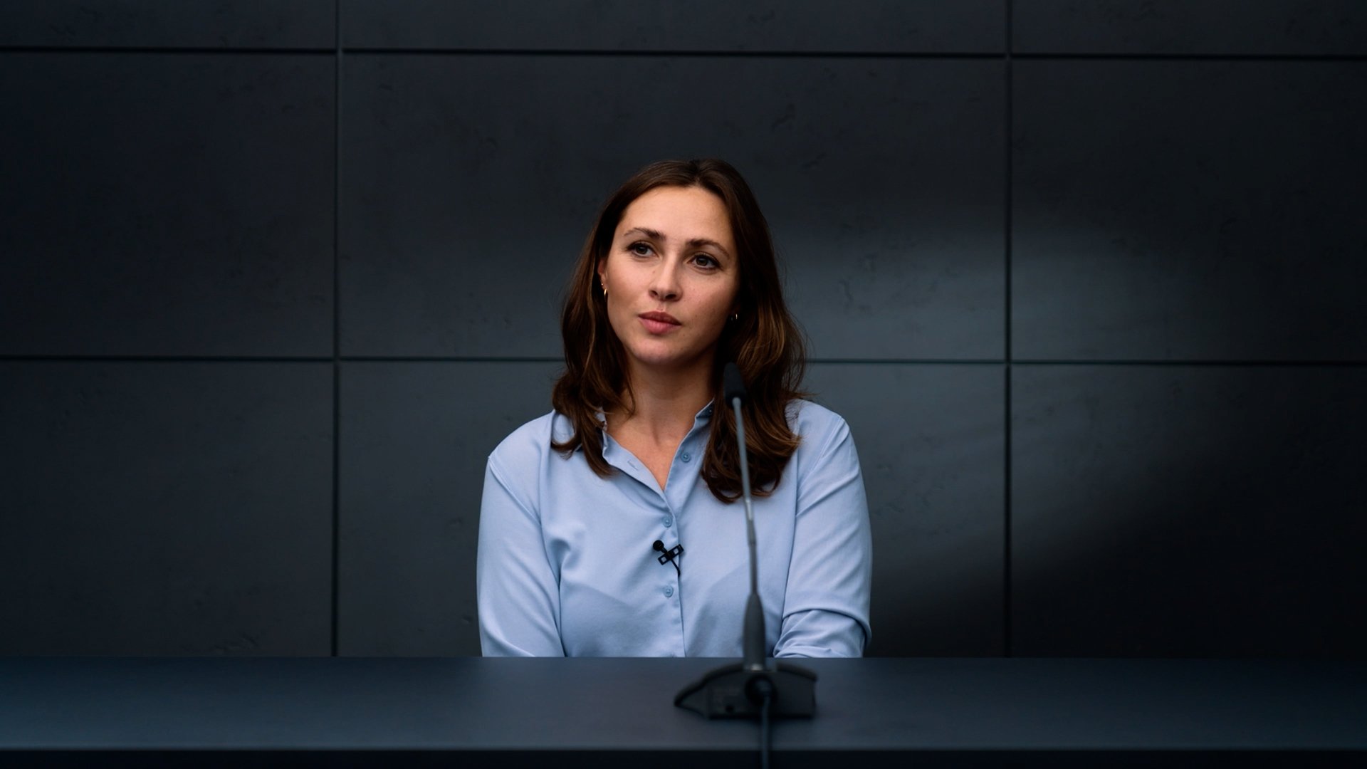 A woman with brown hair in a light blue shirt sits in front of a microphone against a dark gray wall, appearing to be in a formal interview or press conference.