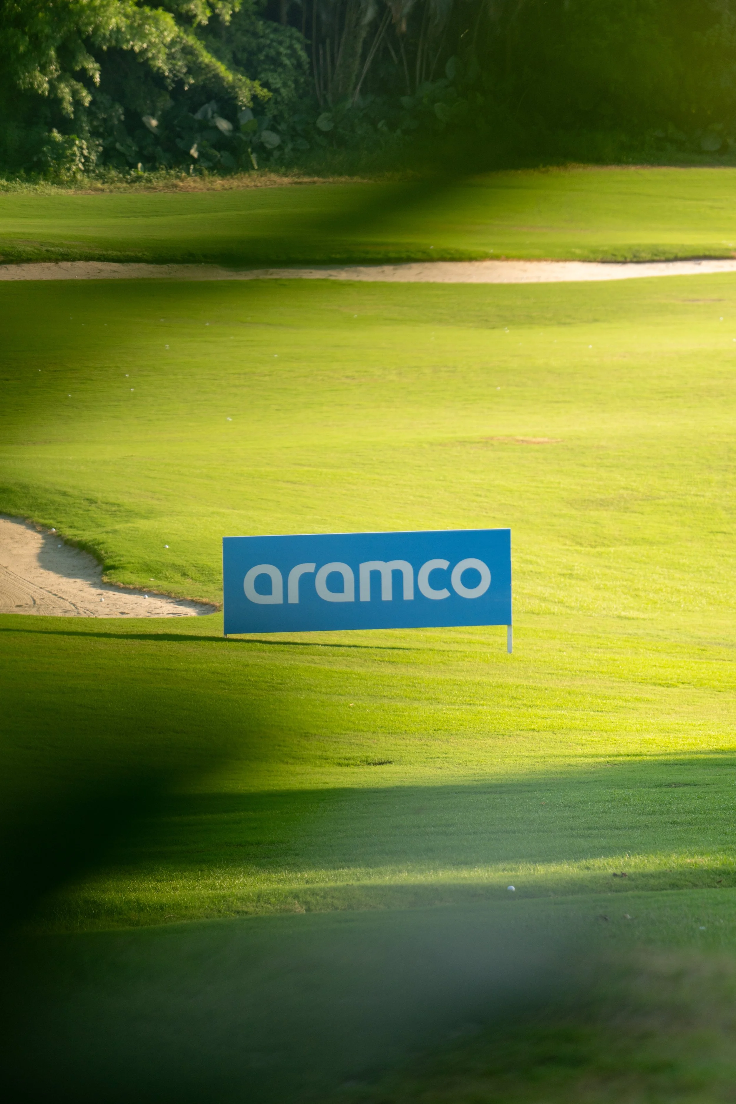A golf course with a blue sign that has the word "aranco" on it, surrounded by green grass and trees in the background.