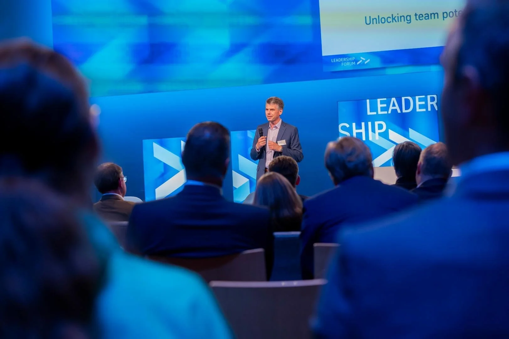A man in a suit giving a presentation at a conference with a blue-themed backdrop that says 'LEADERSHIP'. Audience members in business attire are seated facing him.