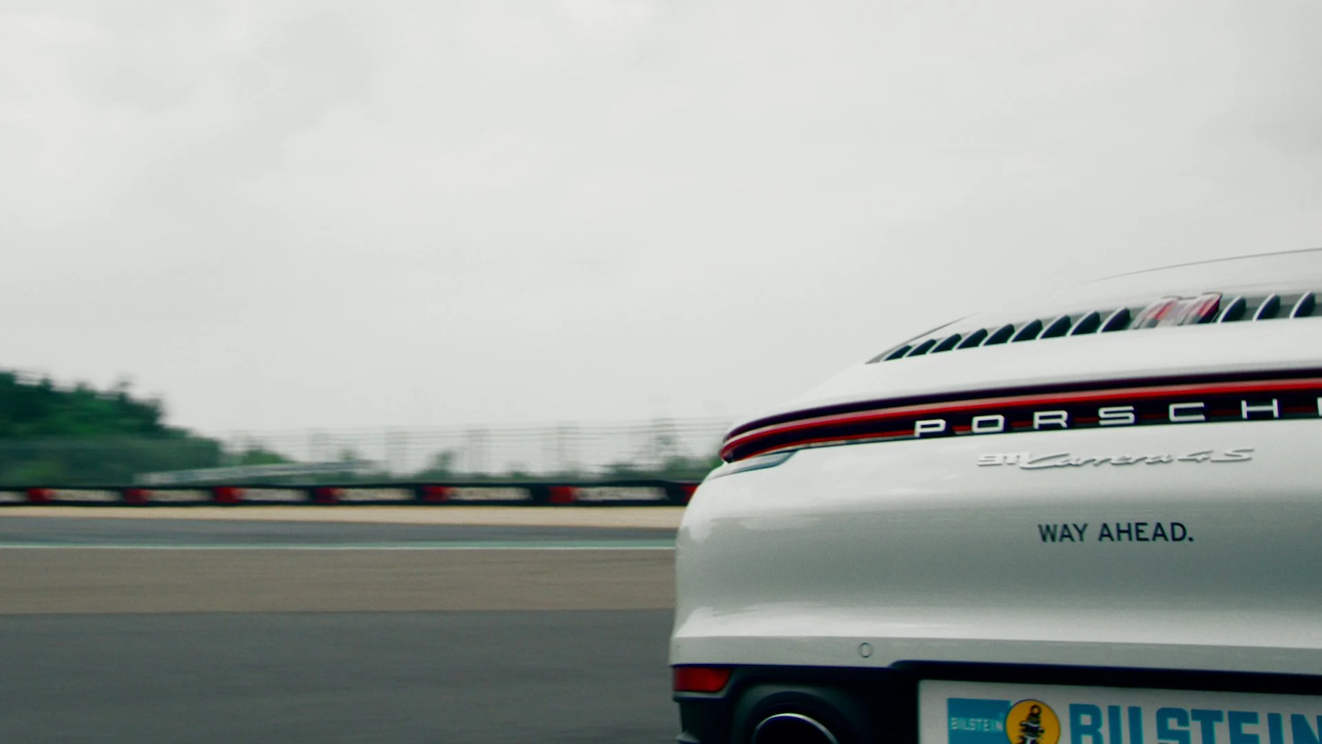 Close-up of the rear of a white Porsche 911 Carrera S sports car on a racetrack, with red and black branding, and the phrase "WAY AHEAD" on the bumper, with blurred track and guardrail in the background.