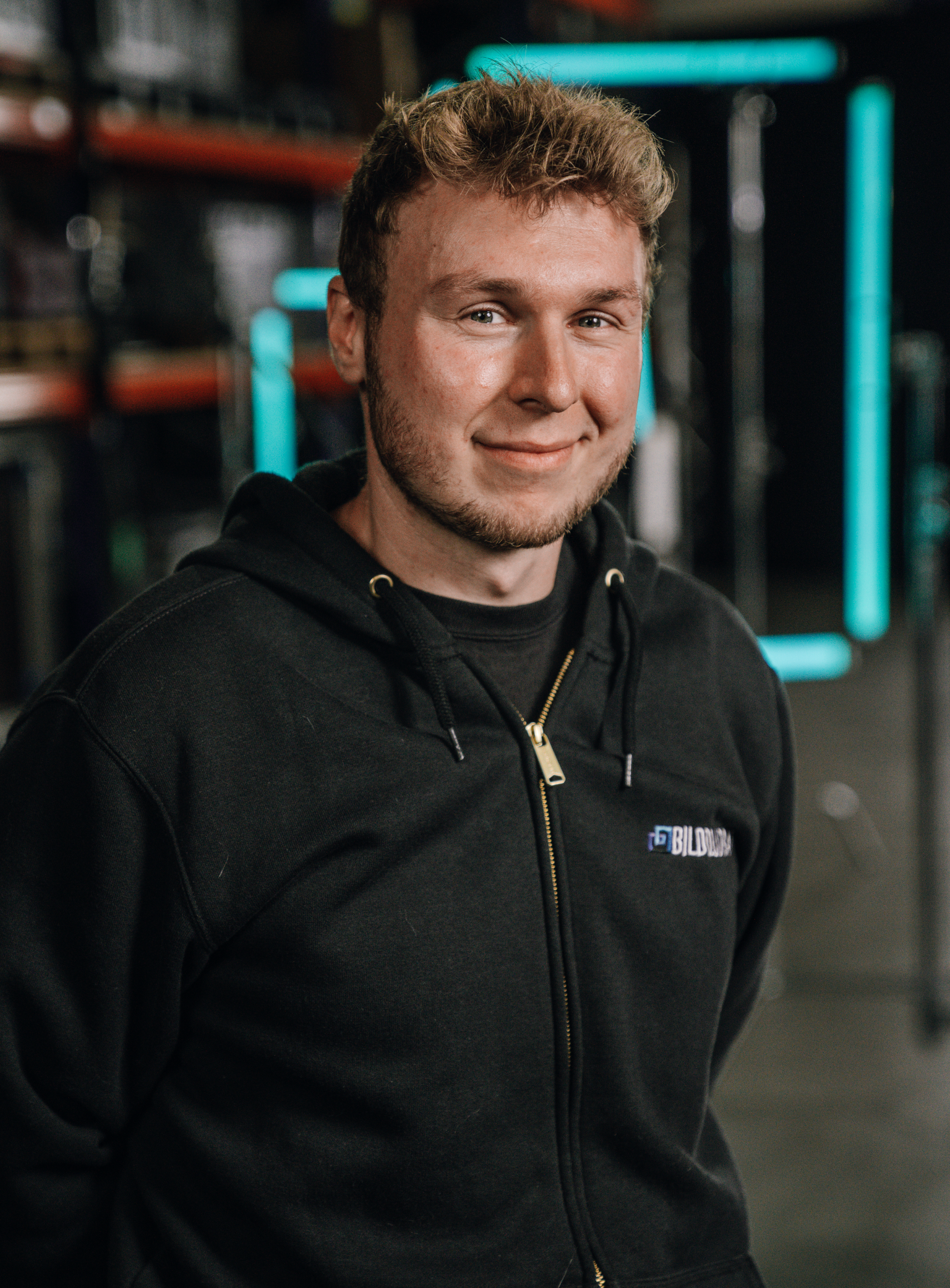 A young man with short, curly hair and a beard, smiling, wearing a black hoodie with a logo, in a gym setting with exercise equipment and neon lights in the background.