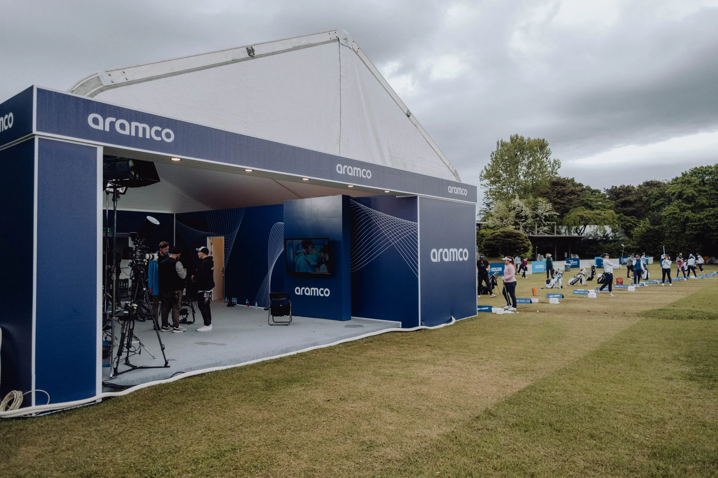 A golf driving range set up with an aramco-branded shelter, with people practicing golf in the open field on a cloudy day, and trees in the background.