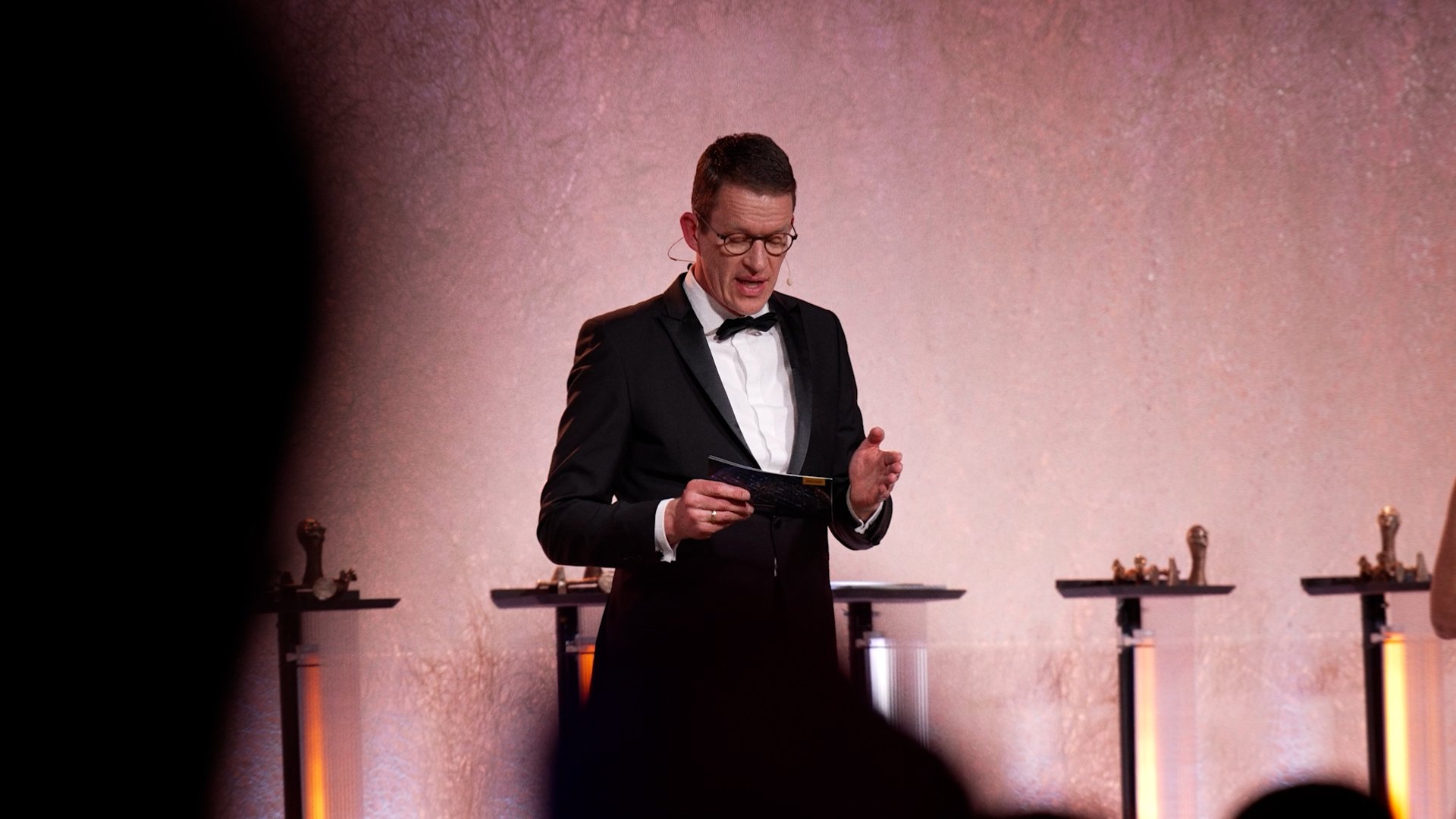 Man in tuxedo standing on stage, reading from a card, with trophies on tables behind him.