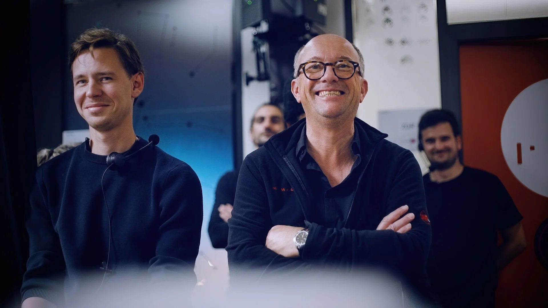 Four men smiling, standing in a room with a dark background and a door on the right. The man in the foreground wears glasses and a dark jacket, with his arms crossed. The man to his left wears a headset around his neck.