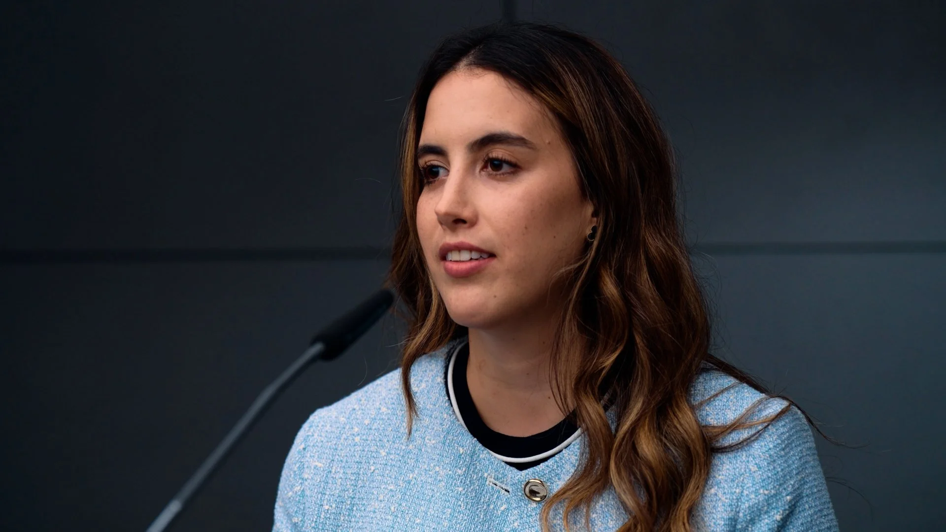 Young woman with wavy brown hair speaking into a microphone at a formal event or conference, wearing a light blue textured blazer with black and white trim.
