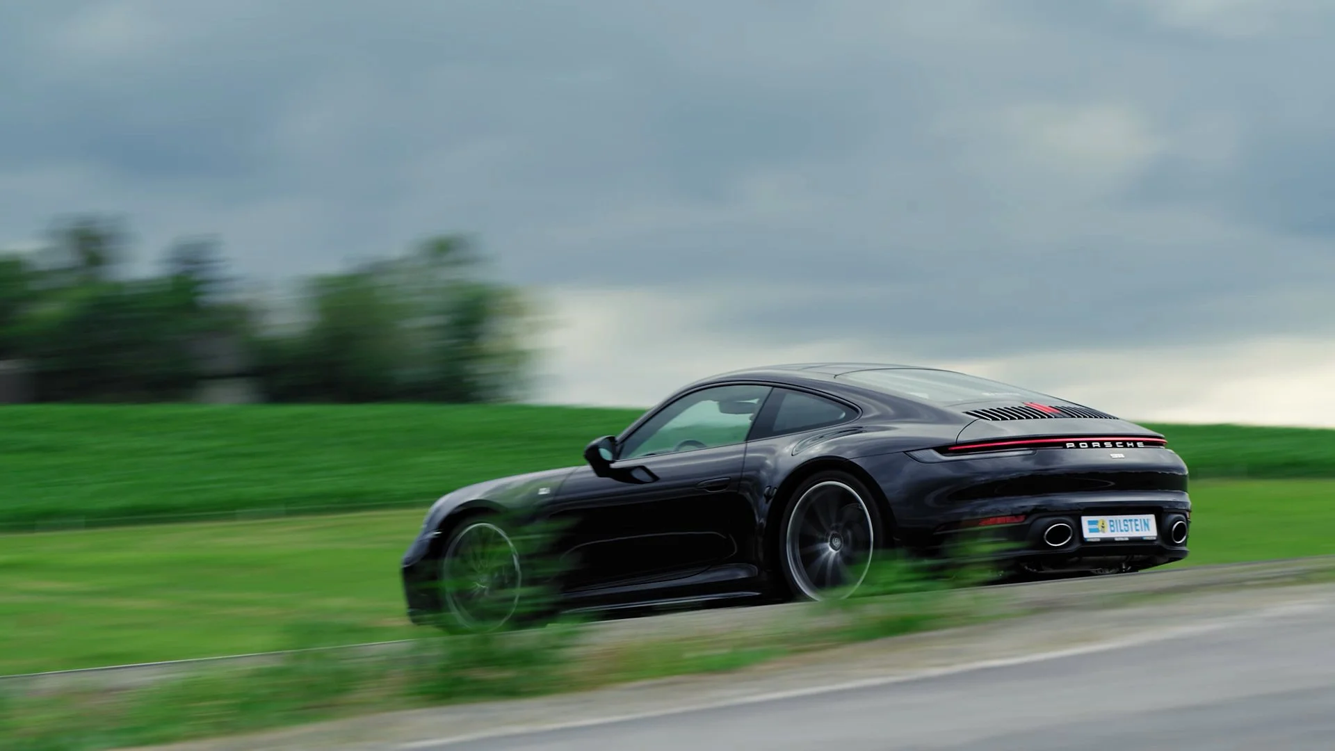 A black Porsche sports car driving on a road with a blurred green field and trees in the background on a cloudy day.