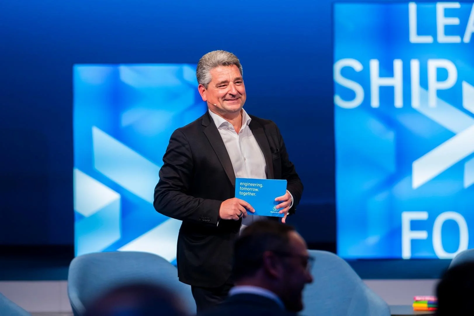 A man in a suit holding a blue booklet at a conference or presentation with a large blue backdrop displaying words related to leadership and engineering.