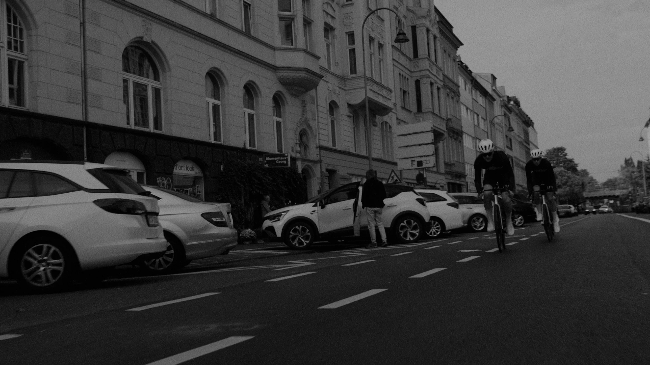Two cyclists riding their bikes on a city street near parked cars and old buildings.