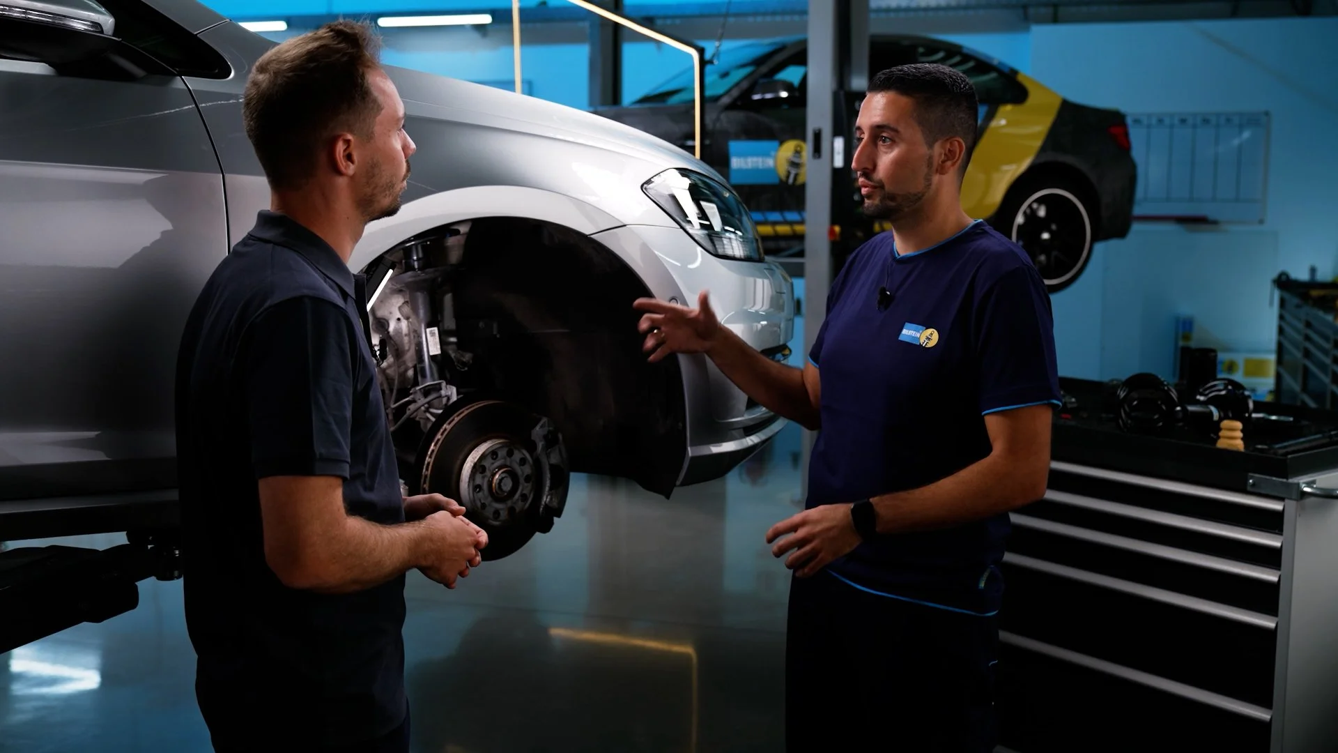 Two mechanics having a discussion in an auto repair shop, near a car lifted on a workbench with the wheel removed, and tools in the background.