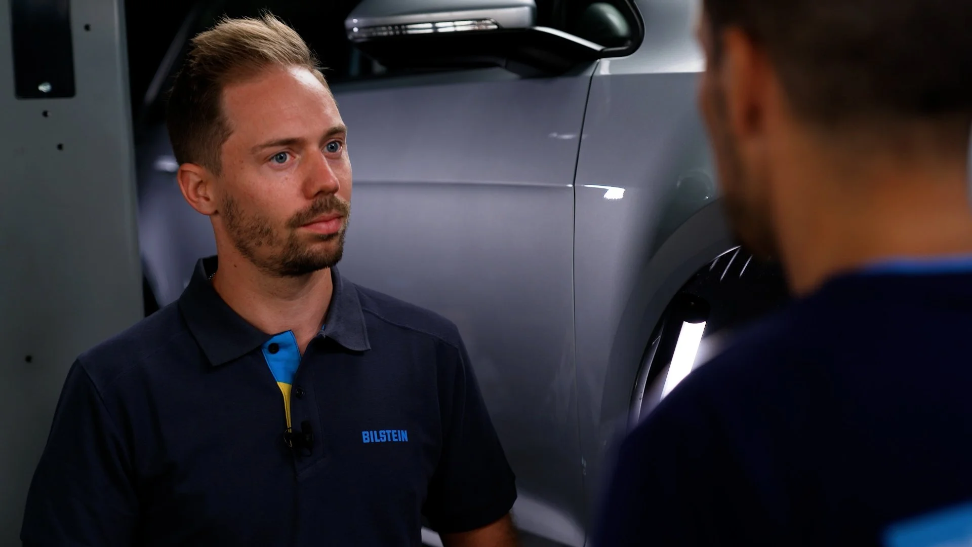Two men in conversation near a gray car, one wearing a Bilstein shirt, inside a workshop or garage.