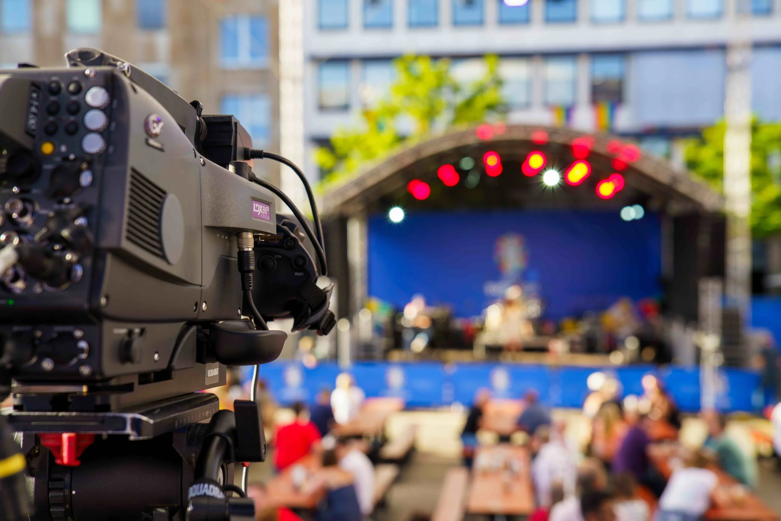 A television camera in focus on the left side, capturing an outdoor stage with blurred audience and performers, during a live event or concert.