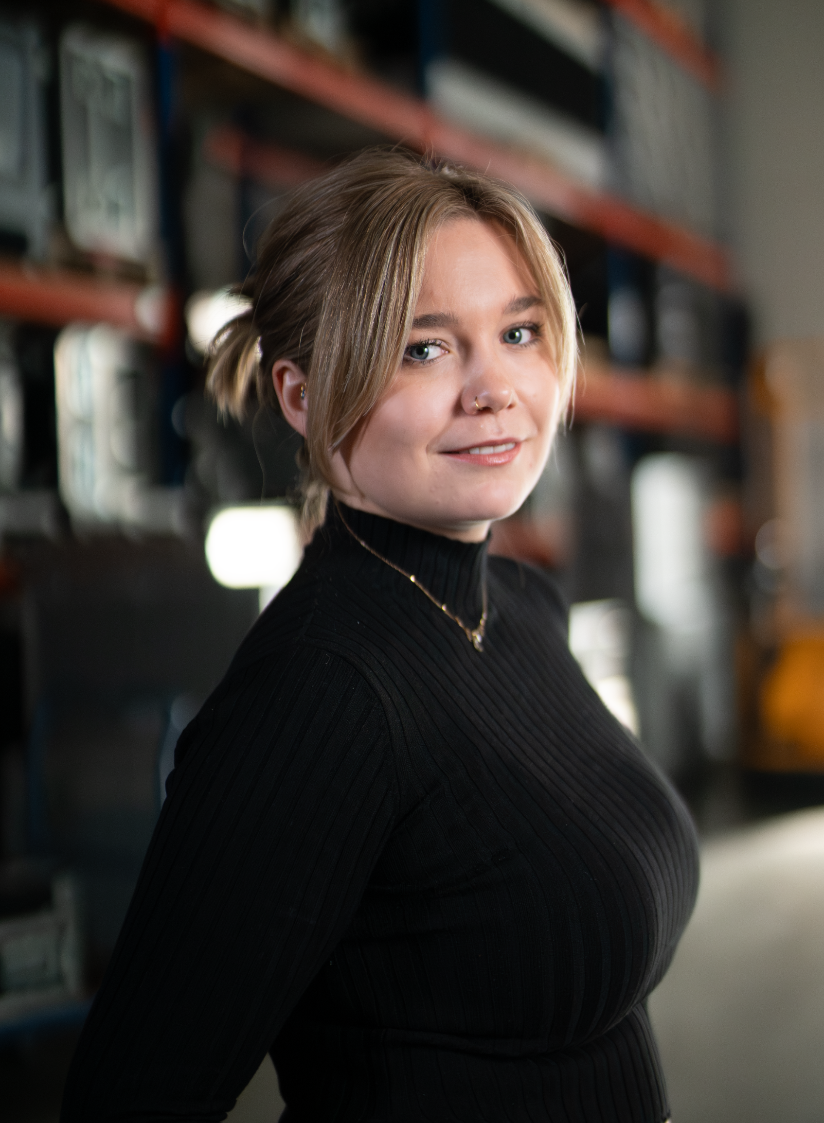 A young woman with blonde hair tied in a loose bun, wearing a black ribbed turtleneck sweater, standing in front of shelves with vintage cameras and equipment, smiling slightly at the camera.