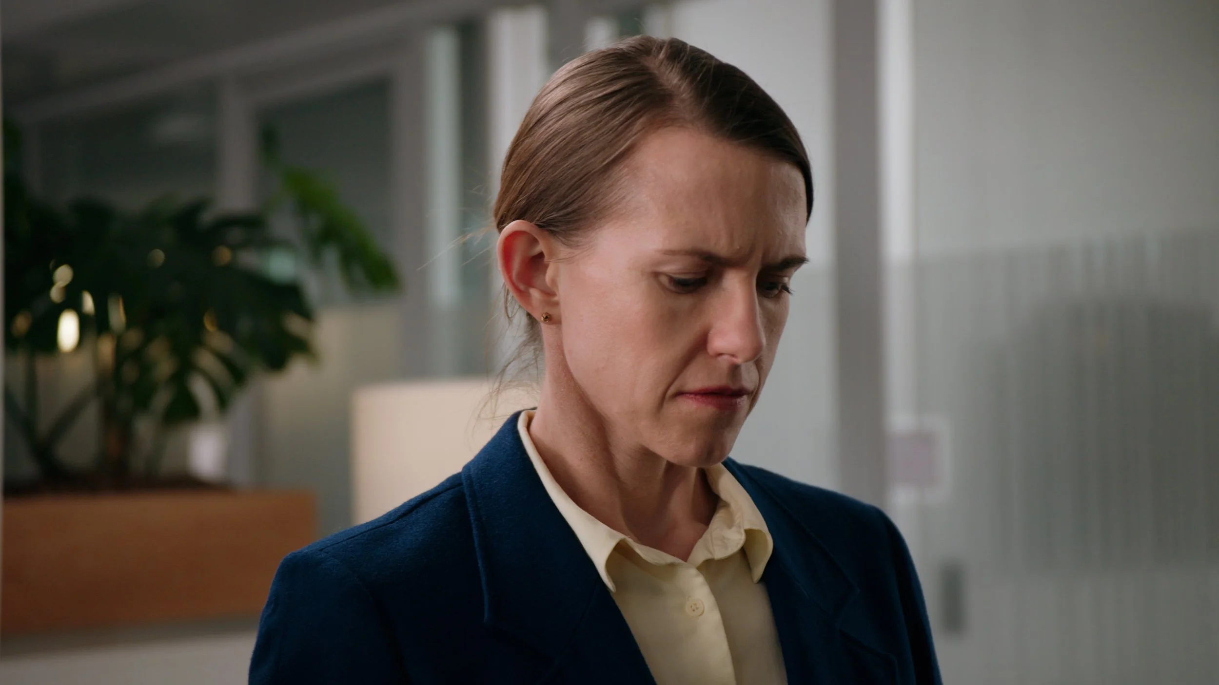 A middle-aged woman with brown hair tied back, wearing a navy blazer and a cream blouse, looks down with a serious expression in a modern office setting.