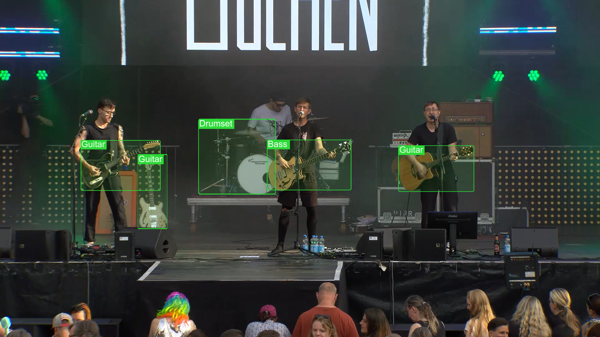 A band performs on stage with a lead singer playing electric guitar, a drummer, and two other guitarists. The audience in front watches the performance.