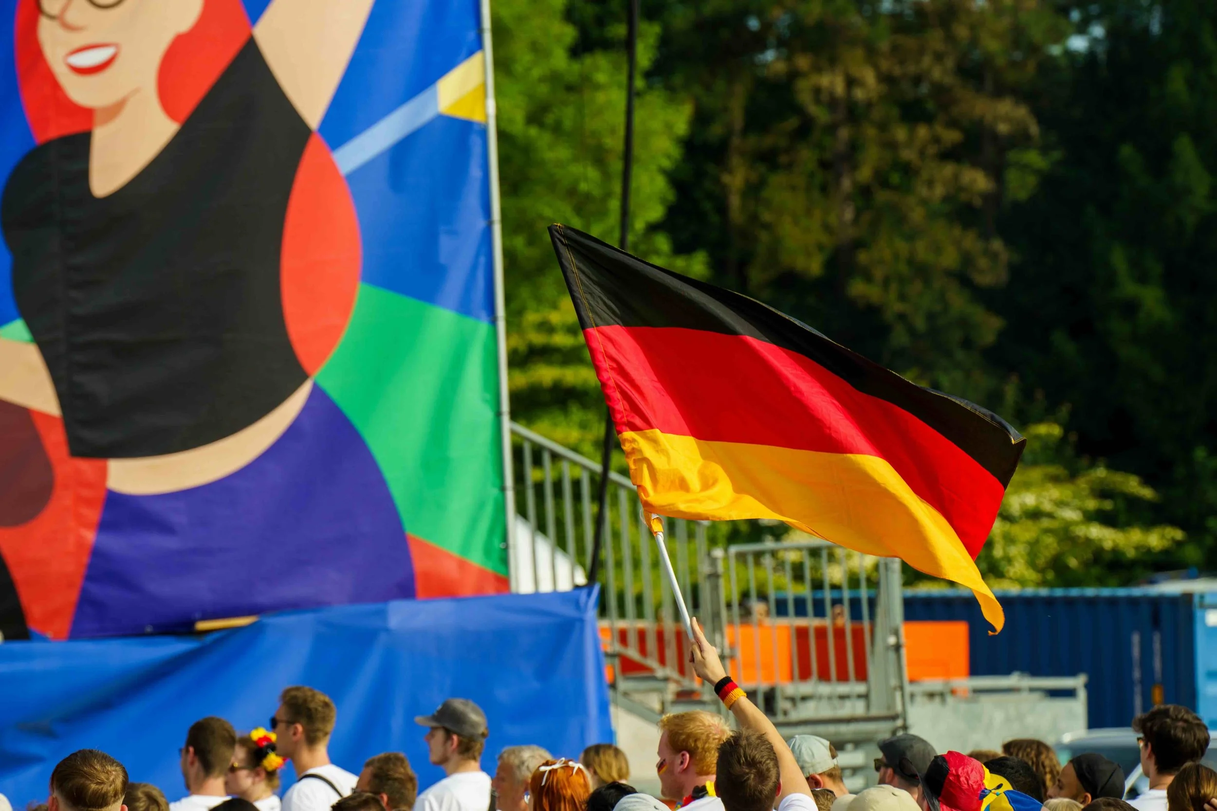 A crowd of people, with some wearing German colors, are gathered outdoors. One person is holding a German flag, and there are large colorful banners or flags in the background, suggesting a celebration or event in Germany.
